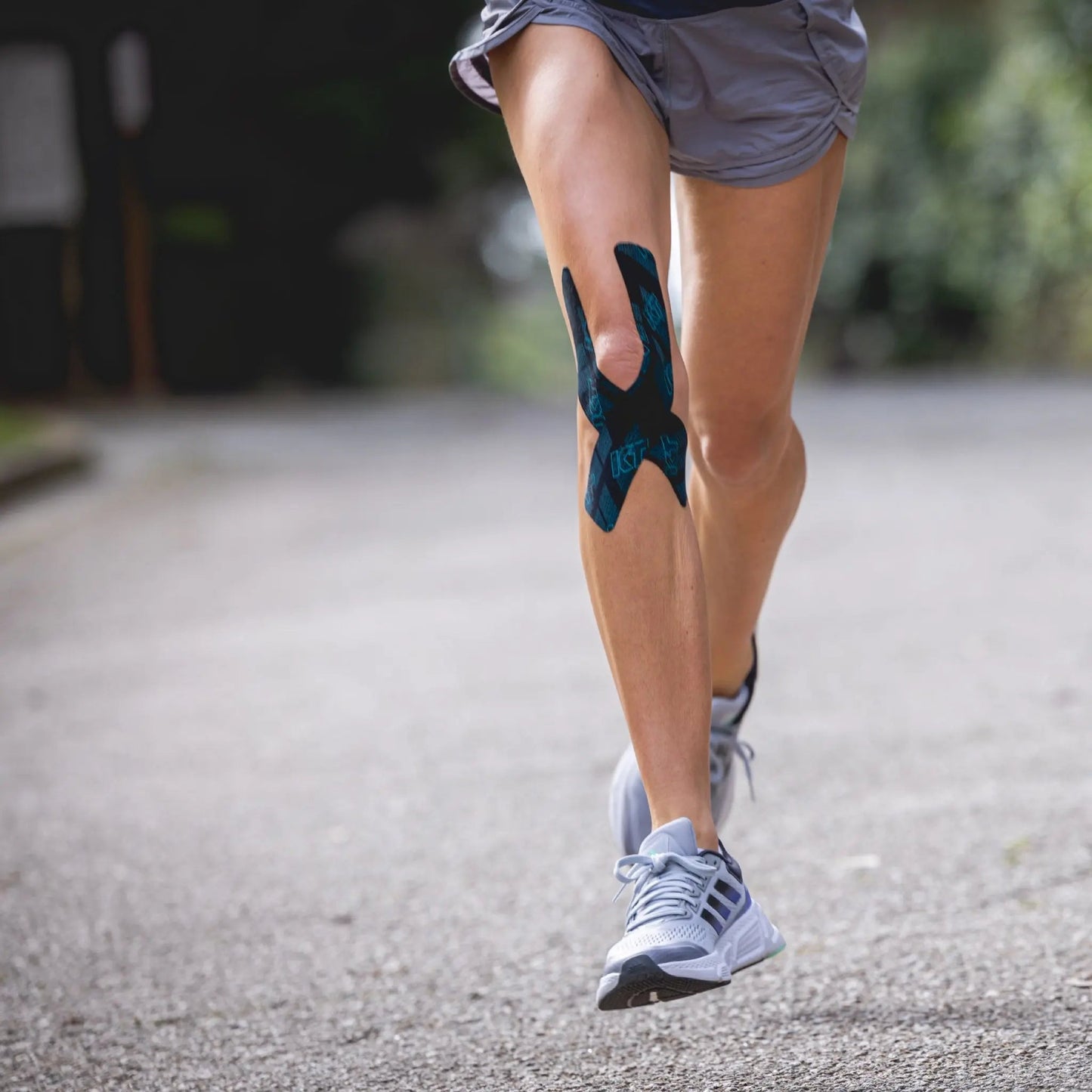 A runner on a paved path wears gray shorts, gray shoes, and KT Tape Pro Ice™ by KT Tape on their right knee for support and pain relief. The focus is on the taped knee, with a blurred background.