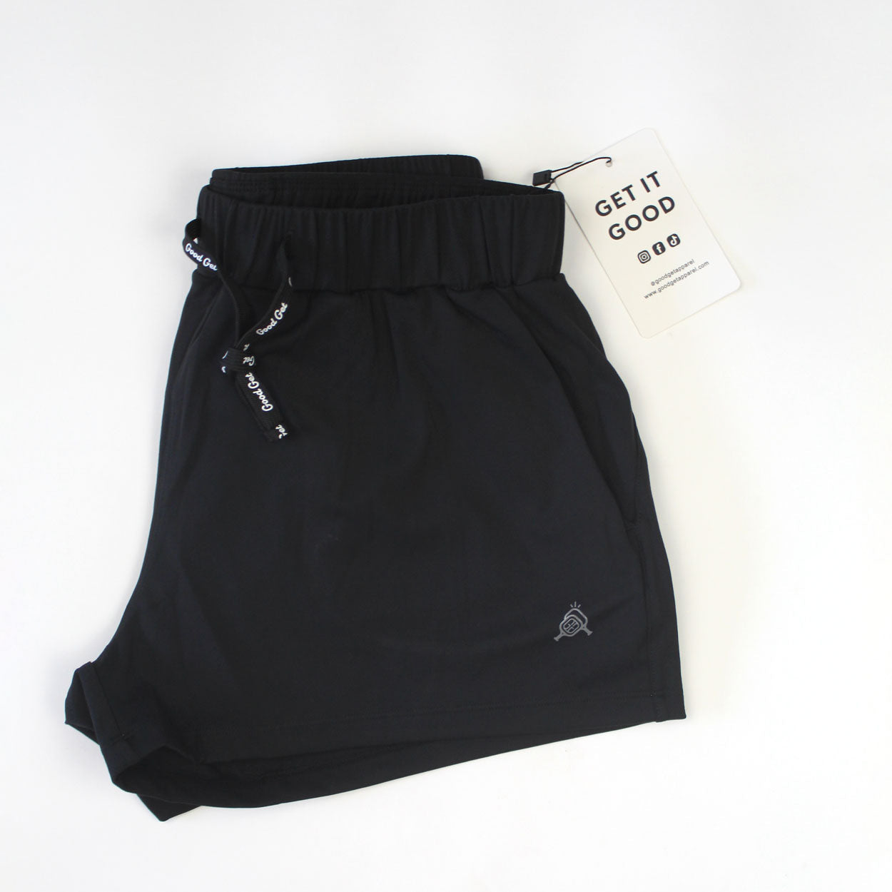 A pair of black Carpe Dink'em Pickleball Shorts by Good Get is neatly folded on a white surface. They feature a drawstring waistband with white logo text, a small embroidered logo near the hem, and a “GET IT GOOD” tag with social media icons.