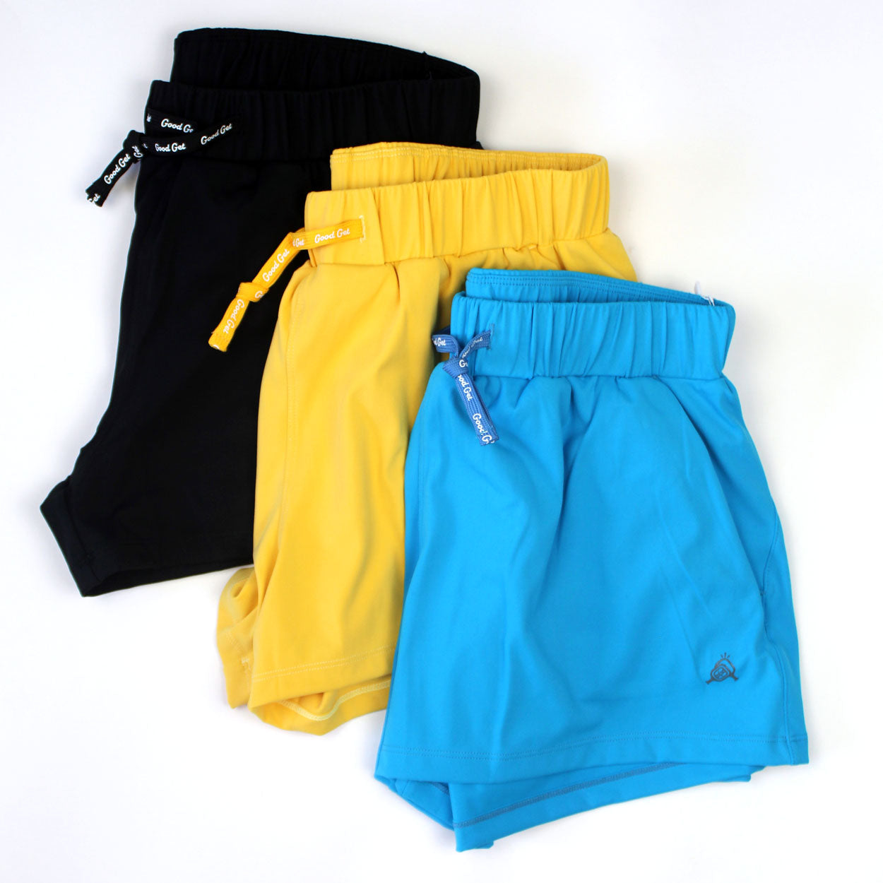Three pairs of Good Get Carpe Dink'em Pickleball Shorts—black, yellow, and bright blue—are fanned out on a white background. Each has an elastic waistband and drawstring; the blue pair displays the Carpe Dink'em logo near the hem.