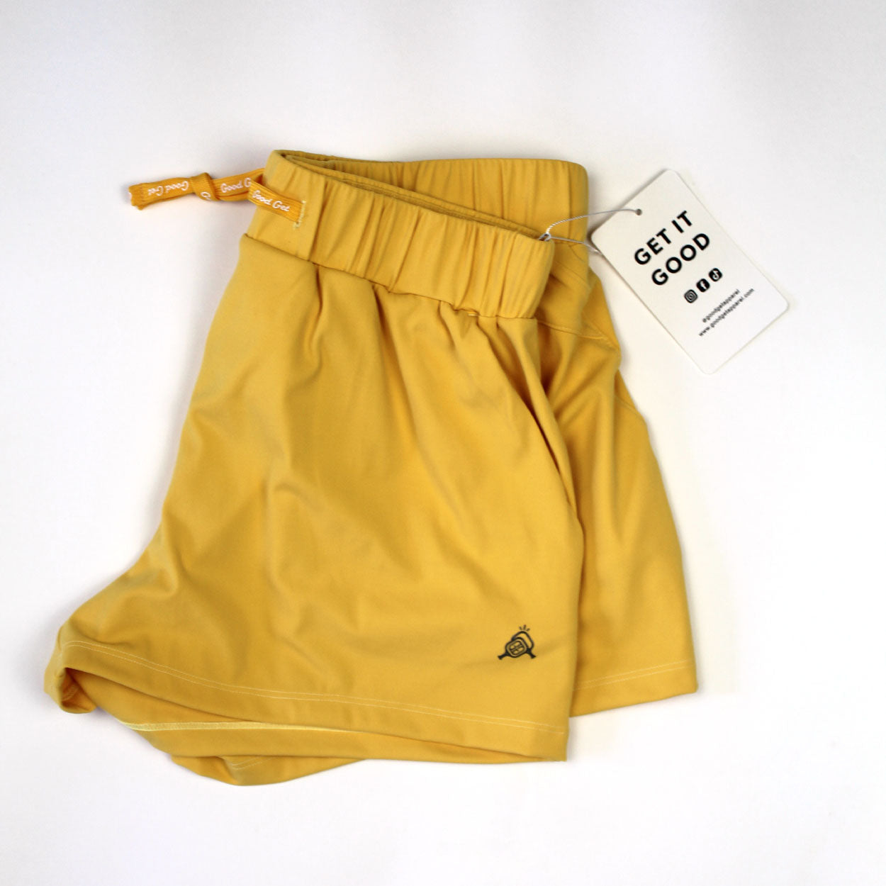 The Carpe Dink'em Pickleball Shorts by Good Get are mustard yellow with an elastic waistband and a small snail logo near the hem. A white tag reads "GET IT GOOD" in bold, capturing their playful spirit. The shorts are shown laid flat on a white background.