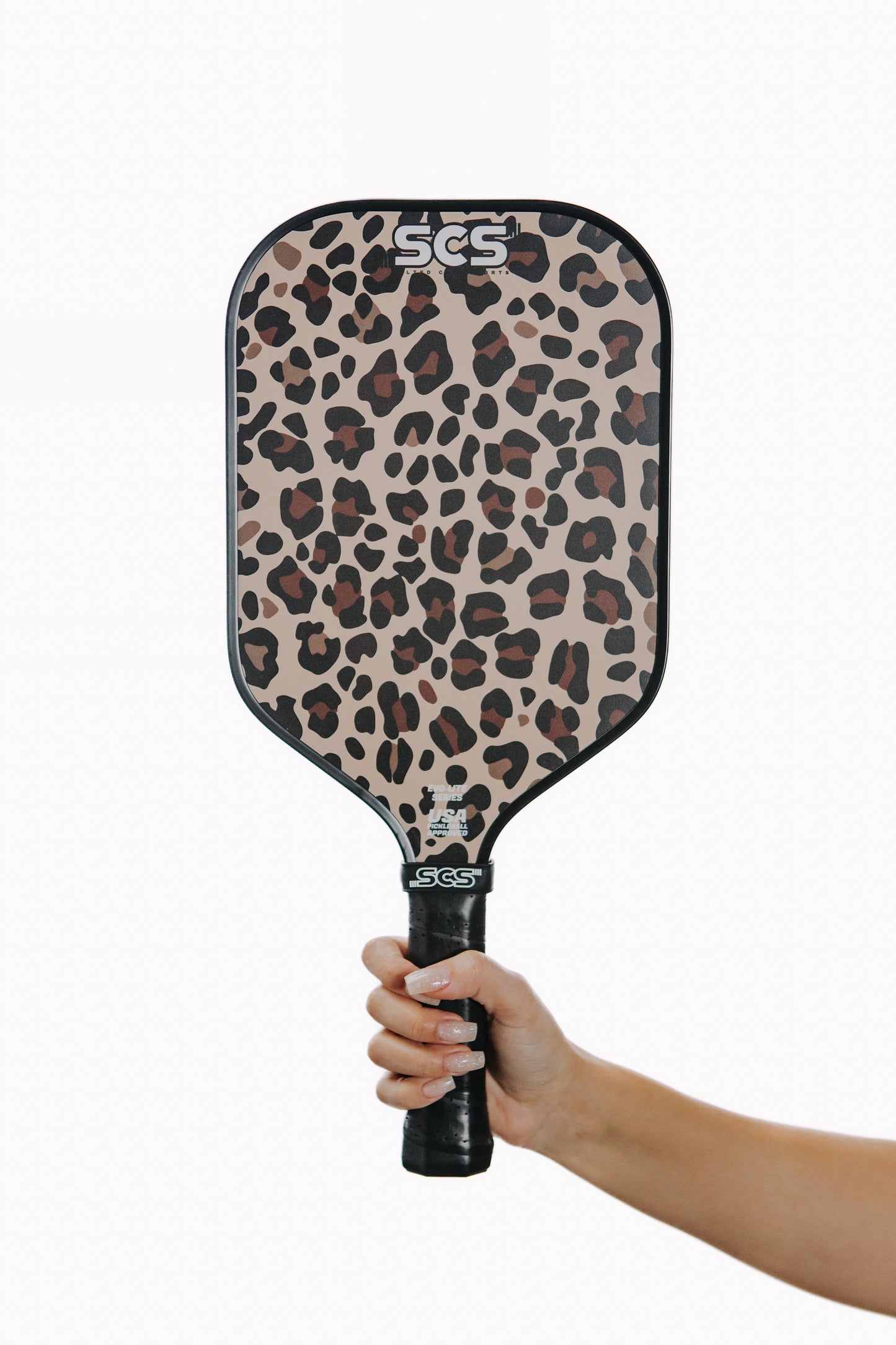 A hand holds the Leopard | Evo-Lite Series pickleball paddle by Salted City Sports, featuring a tan and brown leopard print, "Salted City Sports" near the top, a black grip handle, all set against a plain white background.