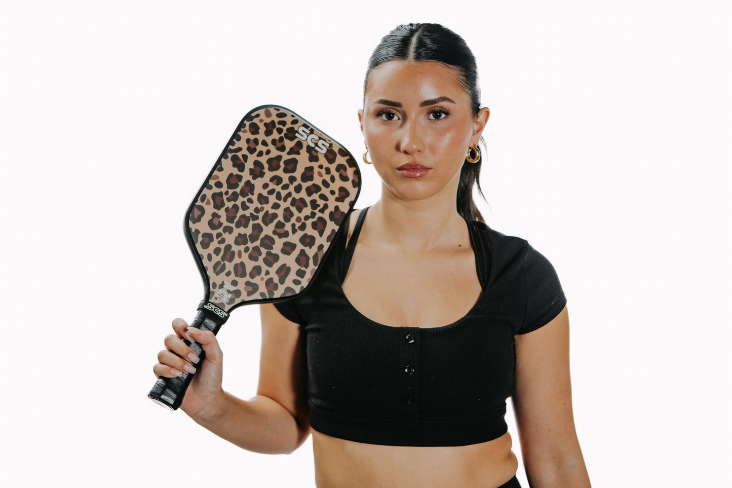 A woman with long dark hair in a ponytail, wearing gold hoop earrings and a black cropped top, holds up the Salted City Sports Leopard | Evo-Lite Series pickleball paddle (USA Pickleball Approved), looking at the camera against a white background.