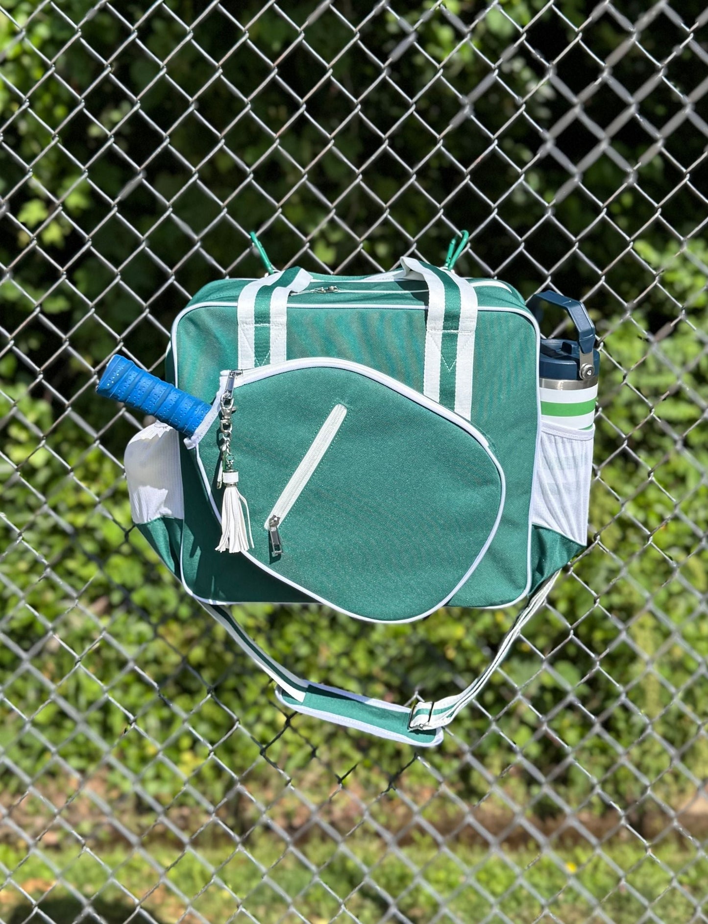 The Fresh Pickle Lola Pickleball Tote Bag in green with white trim hangs on a chain-link fence. Blue-gripped racket and white water bottle sit in side pockets, set against blurred green foliage and sunlight for an outdoor vibe.