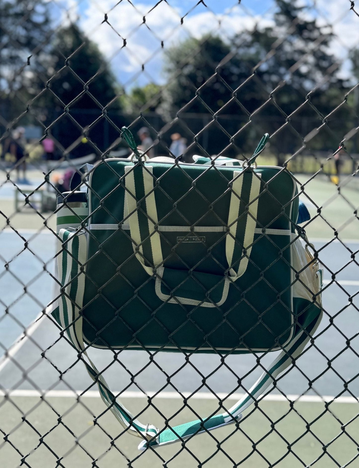 A green and white Fresh Pickle Lola Pickleball Tote Bag hangs by its straps on a black chain-link fence, with people playing tennis on an outdoor court blurred in the background under a blue sky and sunlight casting shadows.