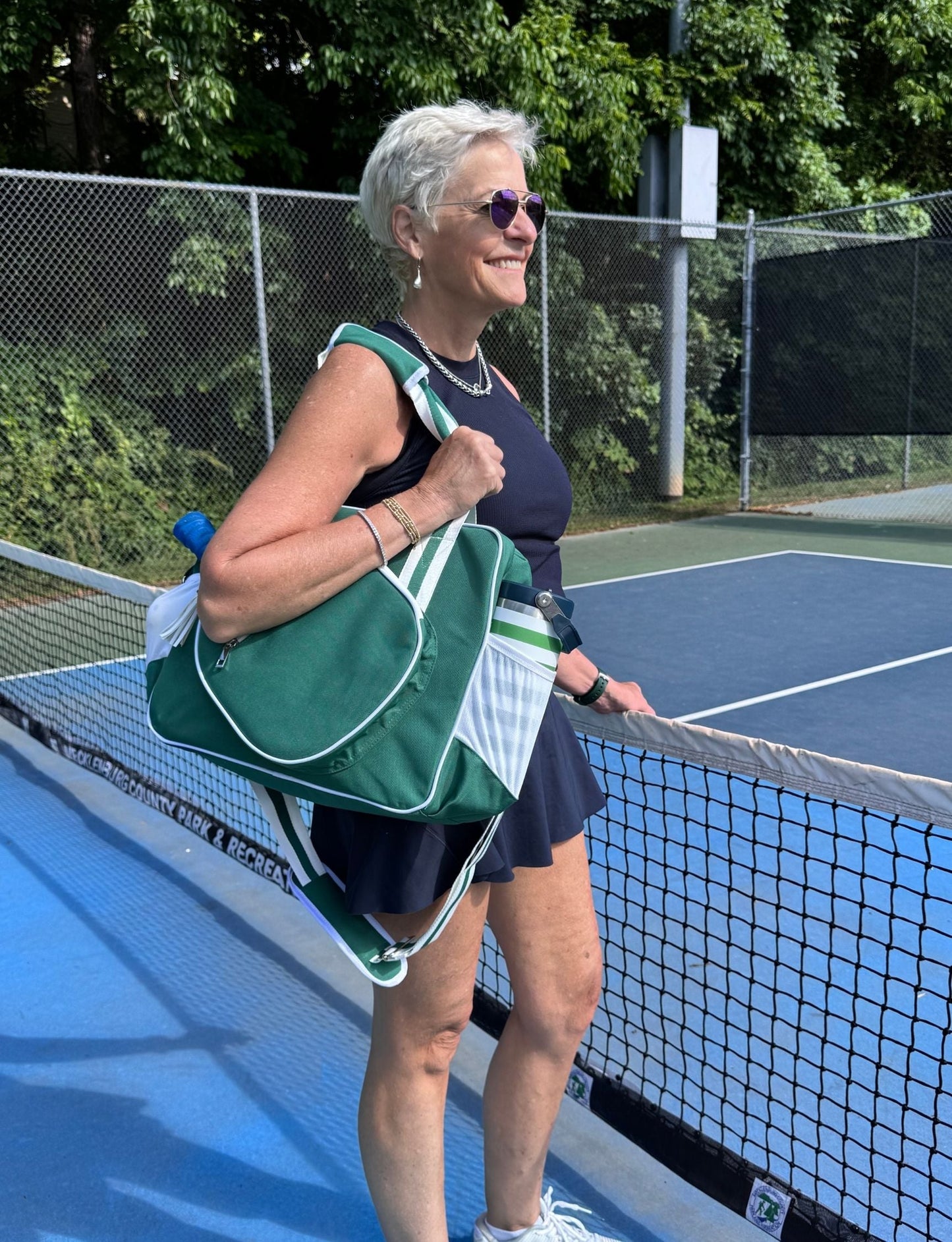 A smiling older woman with short gray hair stands on a tennis court, carrying the Fresh Pickle Lola Pickleball Tote Bag over her shoulder. She wears sunglasses, a navy dress, and white shoes. Green trees and a chain-link fence are in the background.