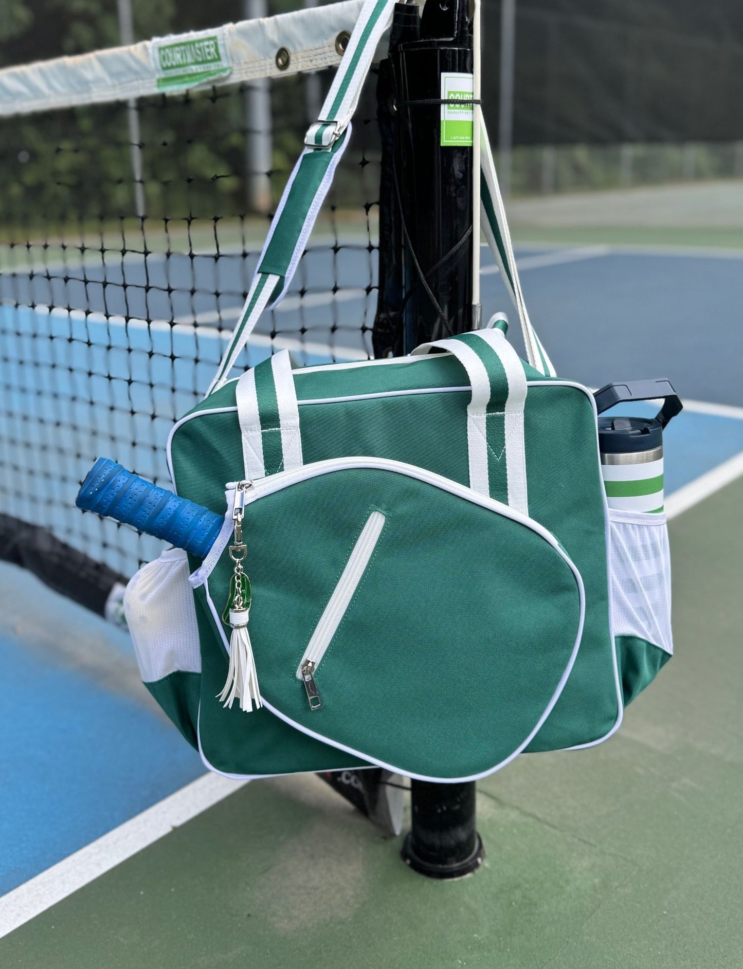 The Fresh Pickle Lola Pickleball Tote Bag in green and white hangs from a tennis net post, holding a blue-handled racket, white tassel keychain, pickleball gear, and a water bottle in its side pocket. Court lines and net are visible behind.