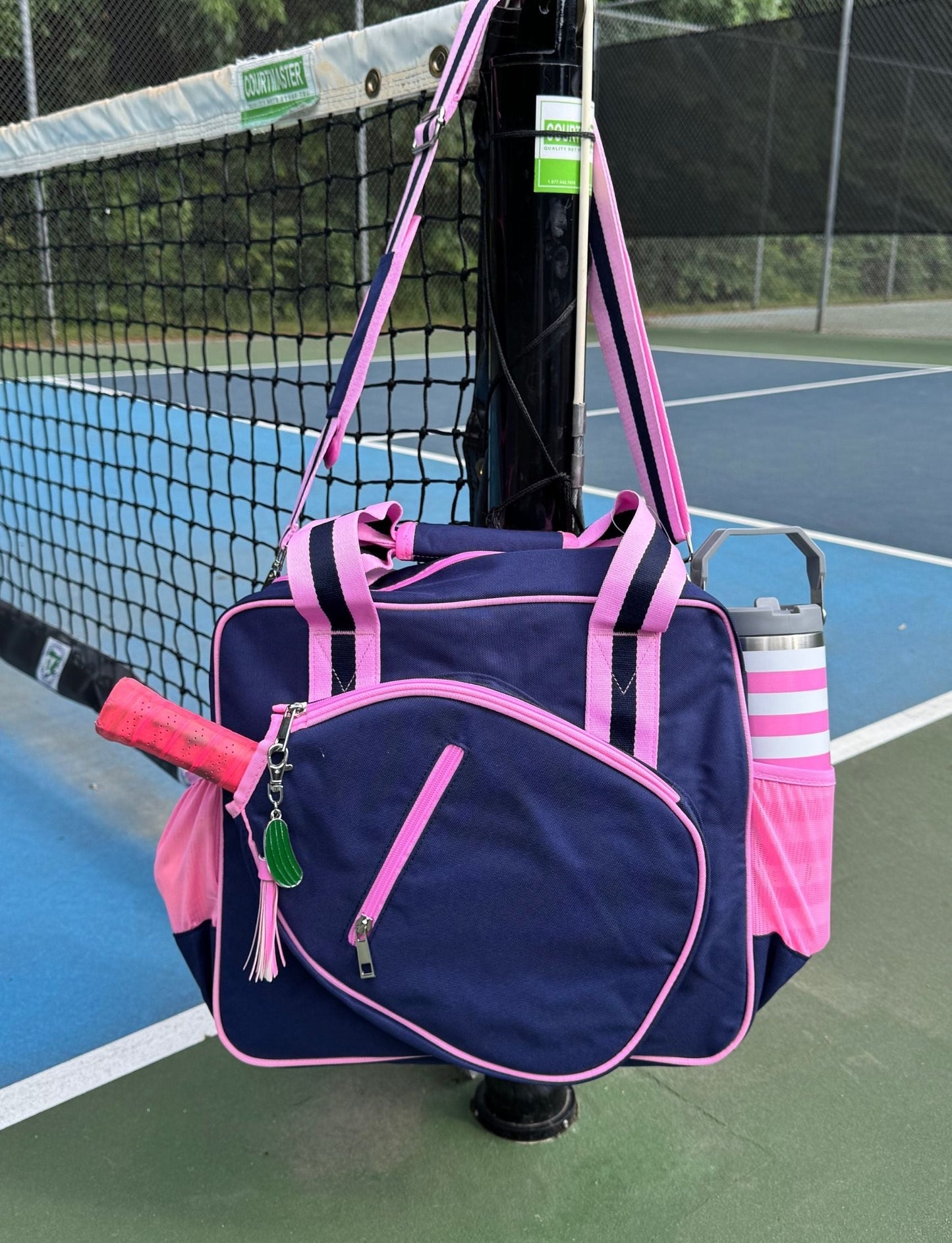 The Fresh Pickle Lola Pickleball Tote Bag in navy blue and pink hangs from a tennis court net post. It features pink straps, a pink-lined front zipper pocket, a side pocket with a pink-striped water bottle, and pickleball accessories.