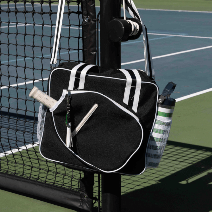 The Fresh Pickle Lola Pickleball Tote Bag, in black with white trim, hangs on a net post. A racket peeks out and a striped water bottle sits in its mesh pocket on a green and blue court with netting behind.