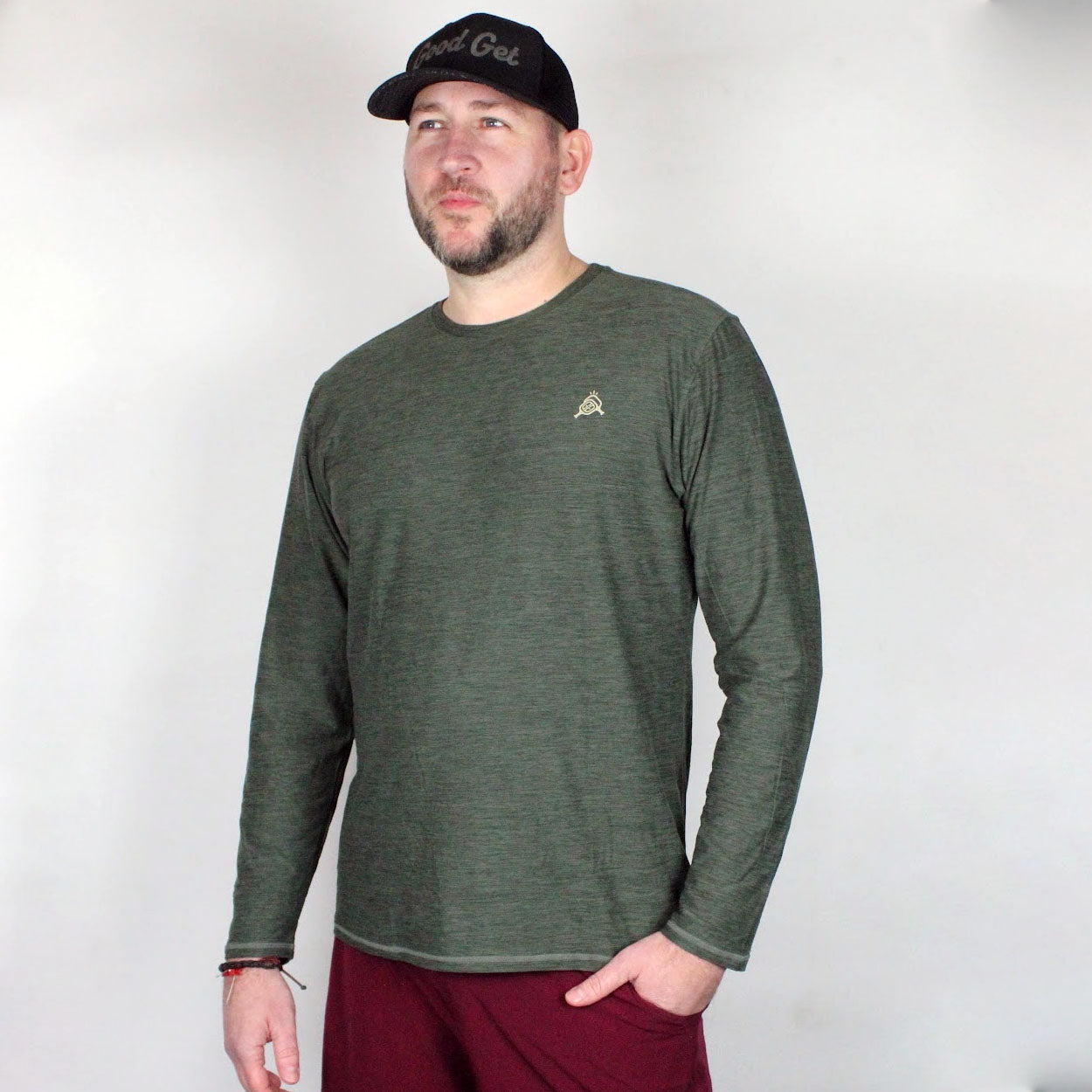 A man with a beard wears a black cap, red pants, and the Good Get AeroBliss Performance Long Sleeve T-shirt in green, featuring a small chest logo. He stands against a light background with one hand in his pocket, looking slightly to the side.