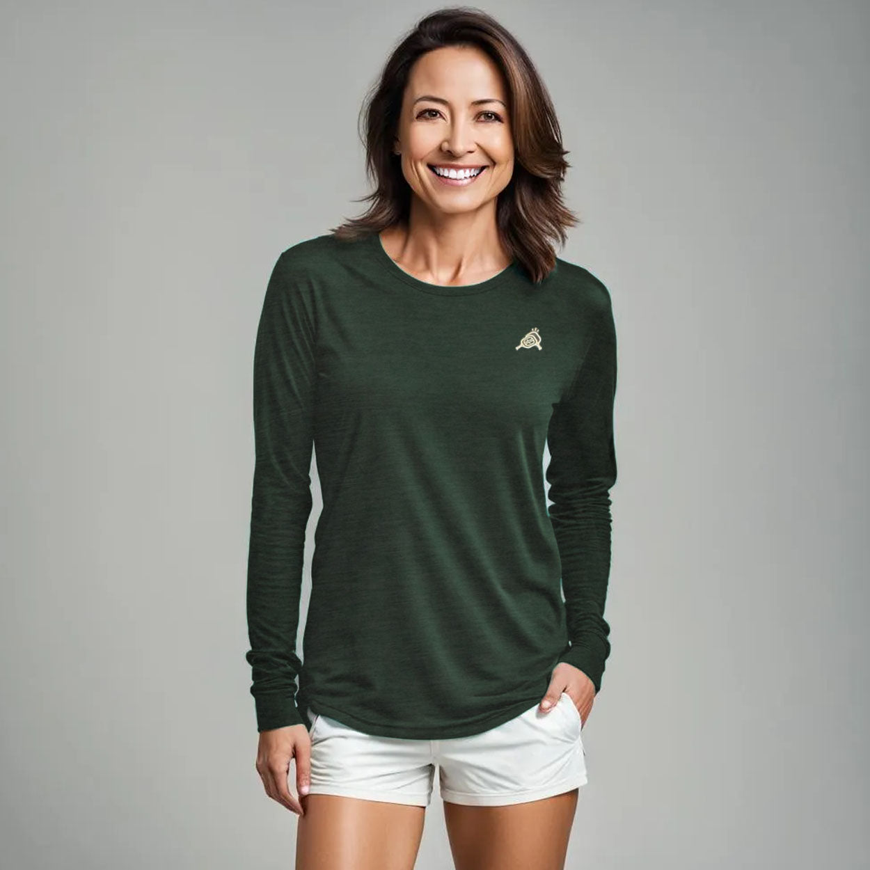 A smiling woman with brown, shoulder-length hair wears the Good Get AeroBliss Performance Long Sleeve T-shirt in dark green, featuring a small emblem on the left chest, and white shorts. She stands against a plain light gray background with her right hand in her pocket.