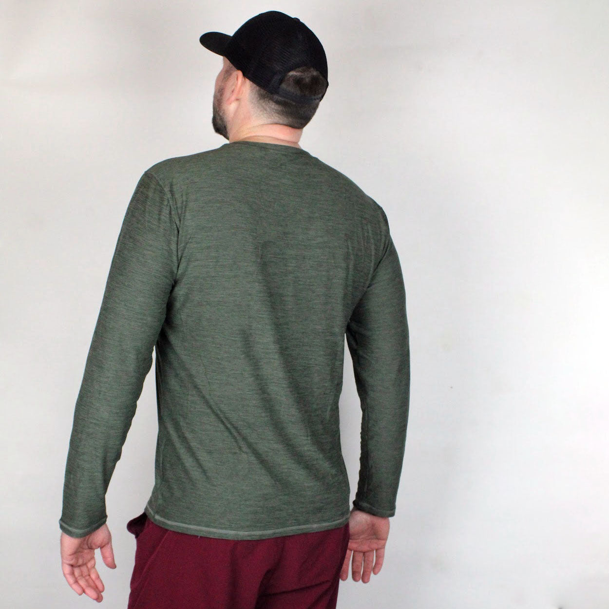 A man stands with his back to the camera, wearing a black baseball cap and the Good Get AeroBliss Performance Long Sleeve T-shirt in olive green, paired with maroon pants. He is against a plain white background with his head slightly turned left.