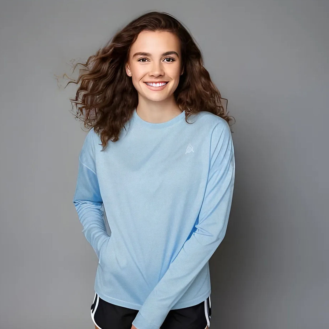 A young woman with wavy brown hair smiles with her hands in her pockets, wearing a Good Get AeroBliss Performance Long Sleeve T-shirt in light blue and black athletic shorts with white trim, standing against a plain gray background.
