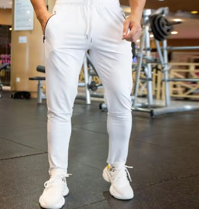 A person stands in a gym wearing DYM Athletics’ Men's Luxury Lounge Jogger | SALE, featuring zipper pockets. White sneakers complete the look, hands in pockets. Weight racks and benches are blurred in the background; upper body is mostly out of frame.