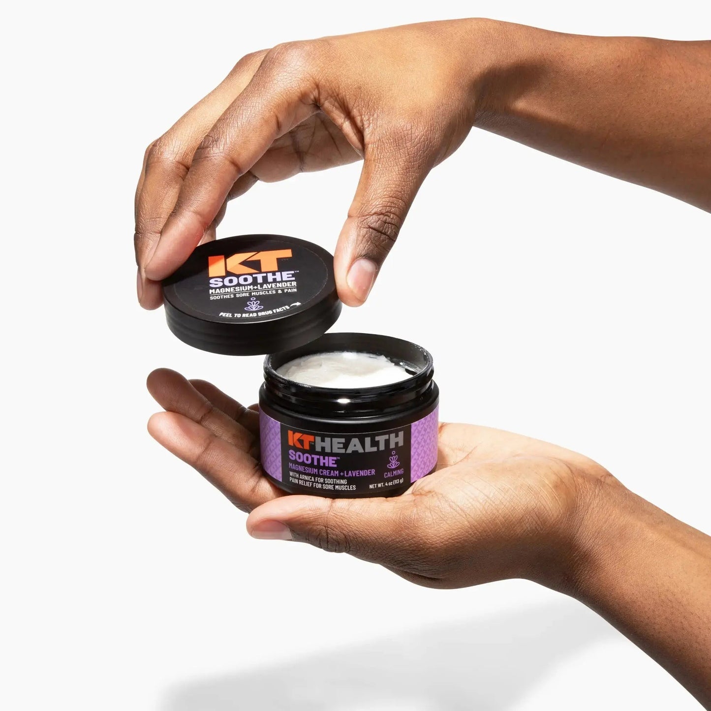 A person opens a black KT Tape jar labeled KT Health Magnesium Creams – Lavender, revealing white cream inside. The purple label with orange and white text stands out against the plain white background.