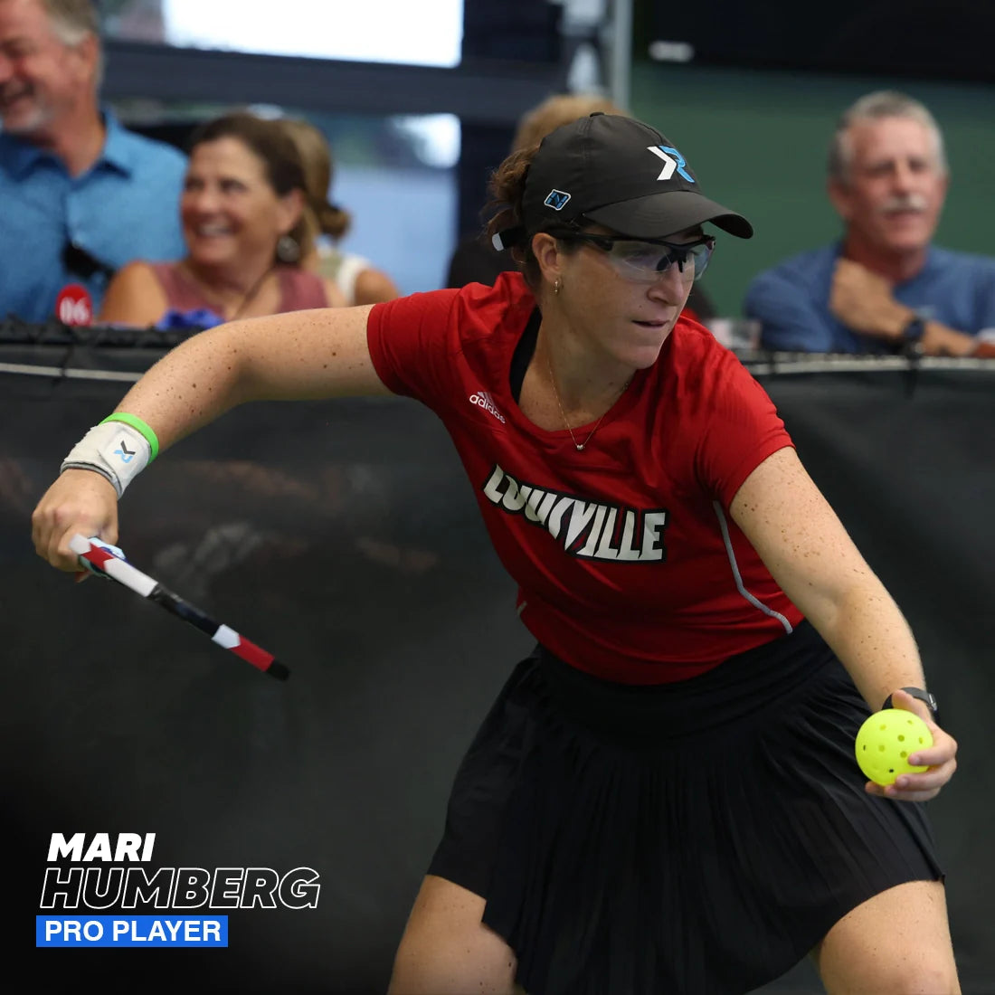 A female pickleball player in a red Louisville shirt and black skirt readies her paddle, equipped with a pink Hesacore Grip Pickleball Grip (5.25" long, regular feel), to hit a yellow ball. She wears glasses, cap, wristband. "Mari Humberg Pro Player" appears left.