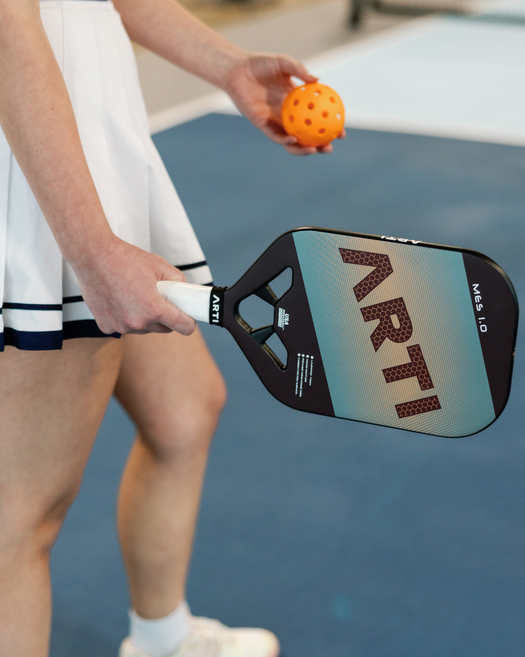 A person in a white tennis skirt holds an ARTI Carbon Fiber "MASTERY ELITE 1.0" paddle by ARTI in one hand and an orange pickleball in the other while standing on a blue indoor court. Only their lower body and hands are visible.
