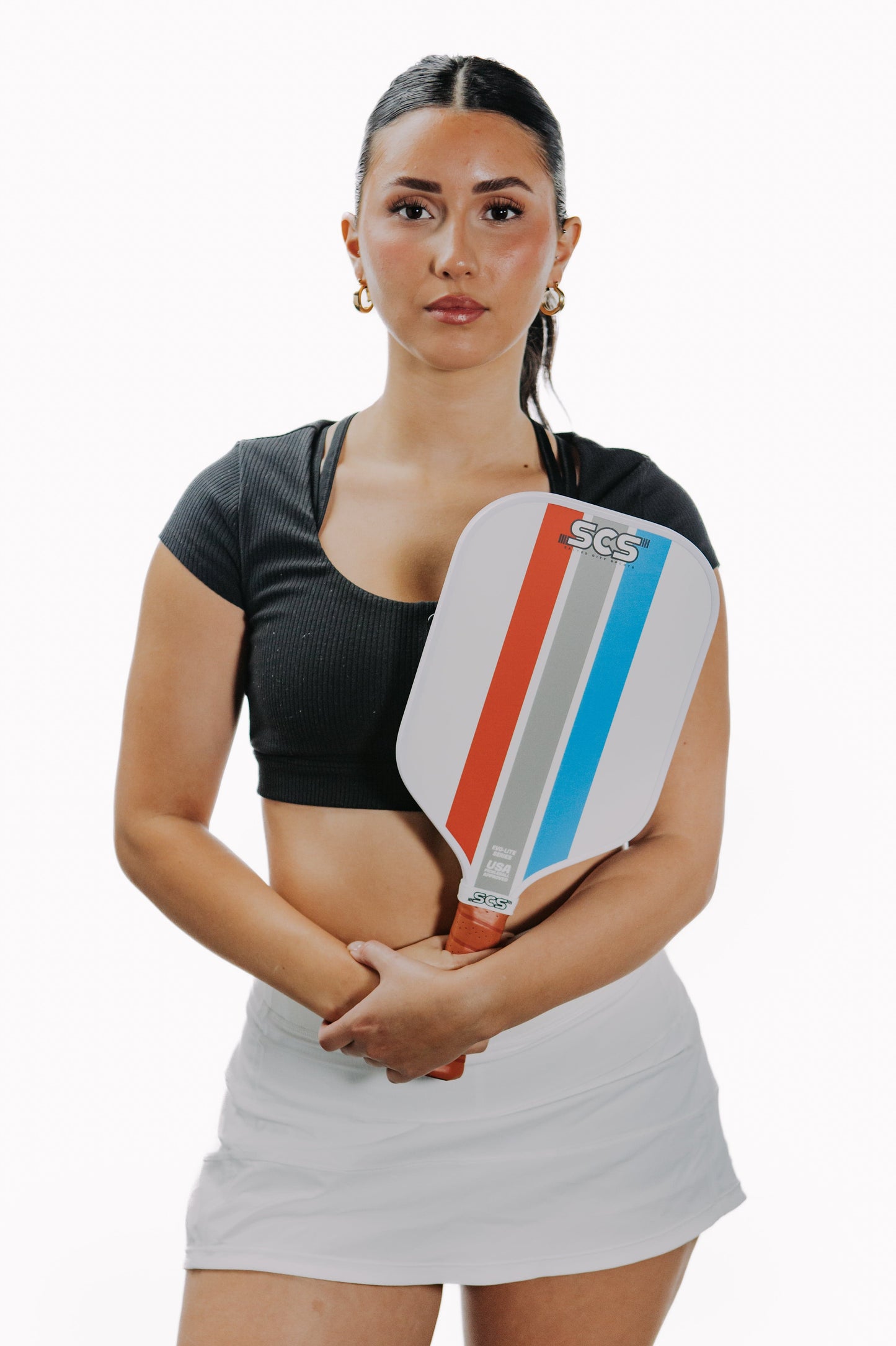 A woman with dark hair in a ponytail, dressed in a black crop top and white skirt, stands facing forward holding a Salted City Sports Mav | Evo-Lite Series pickleball paddle with polypropylene core against a white background.