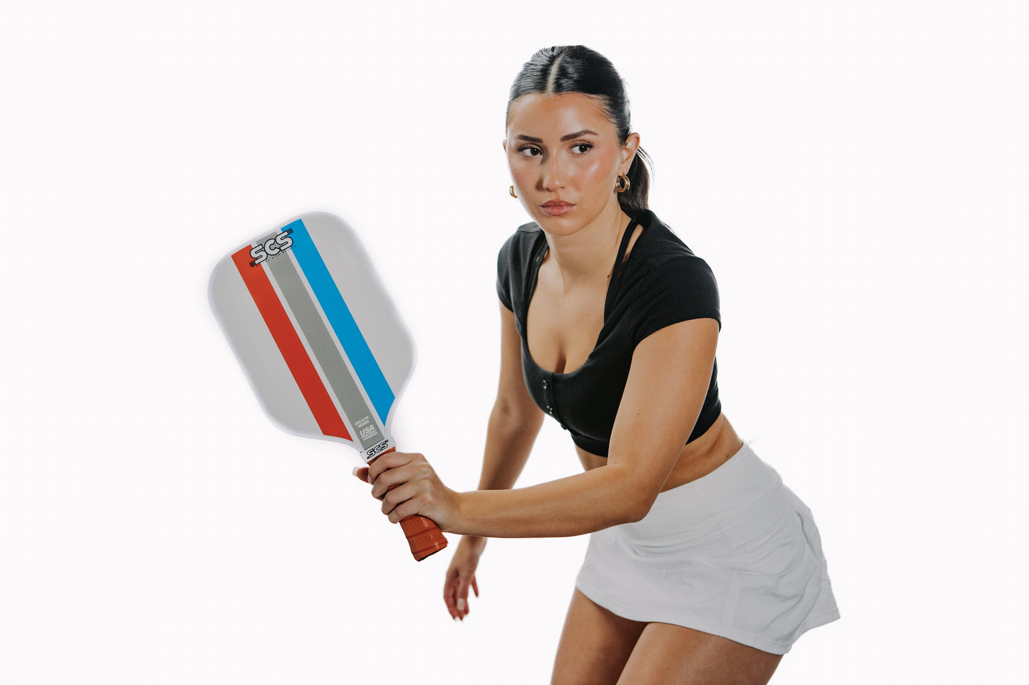 A woman with dark hair in a ponytail, sporting a black crop top and white skirt, stands ready with the Salted City Sports Mav | Evo-Lite Series pickleball paddle (polypropylene core) against a plain white background. She appears focused and prepared to play.