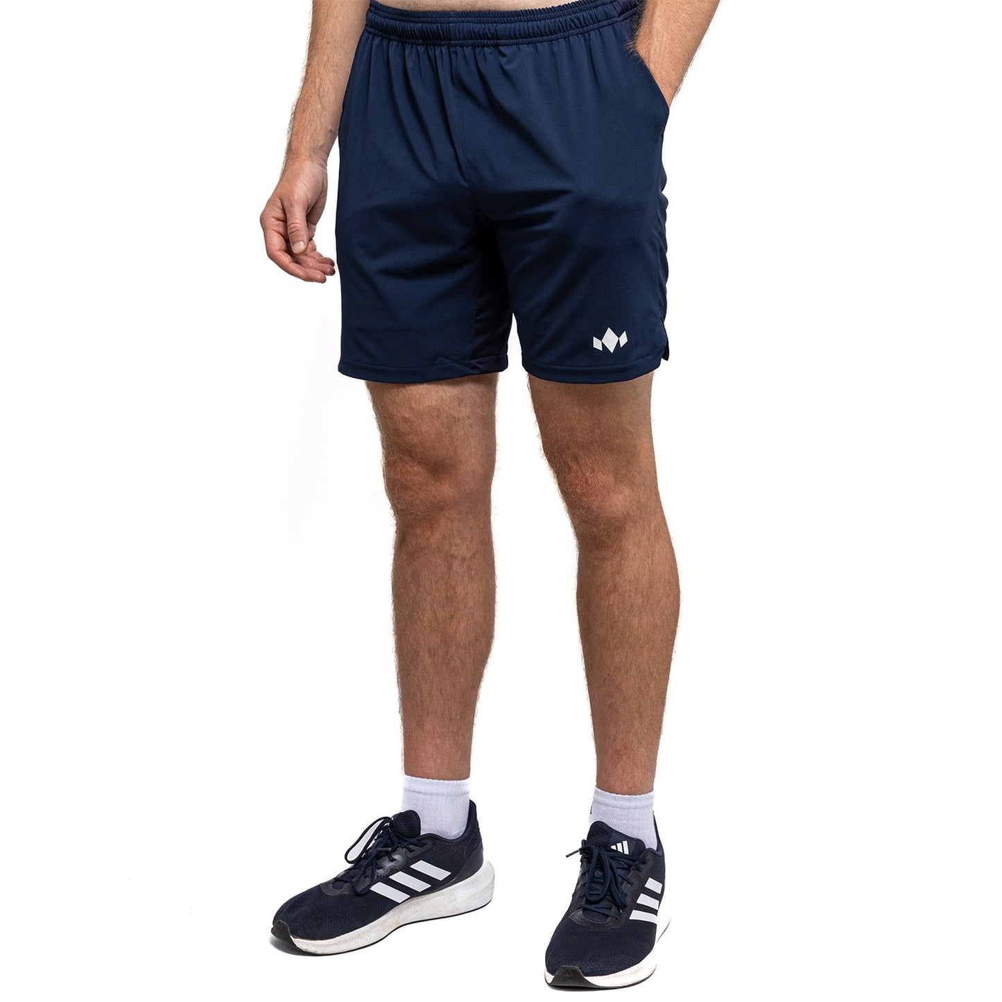A person stands with one hand in a pocket, wearing Diadem Sports Men's Essential 8" Short in navy featuring a small white logo on the left leg, paired with white socks and navy Adidas sneakers, against a plain white background.