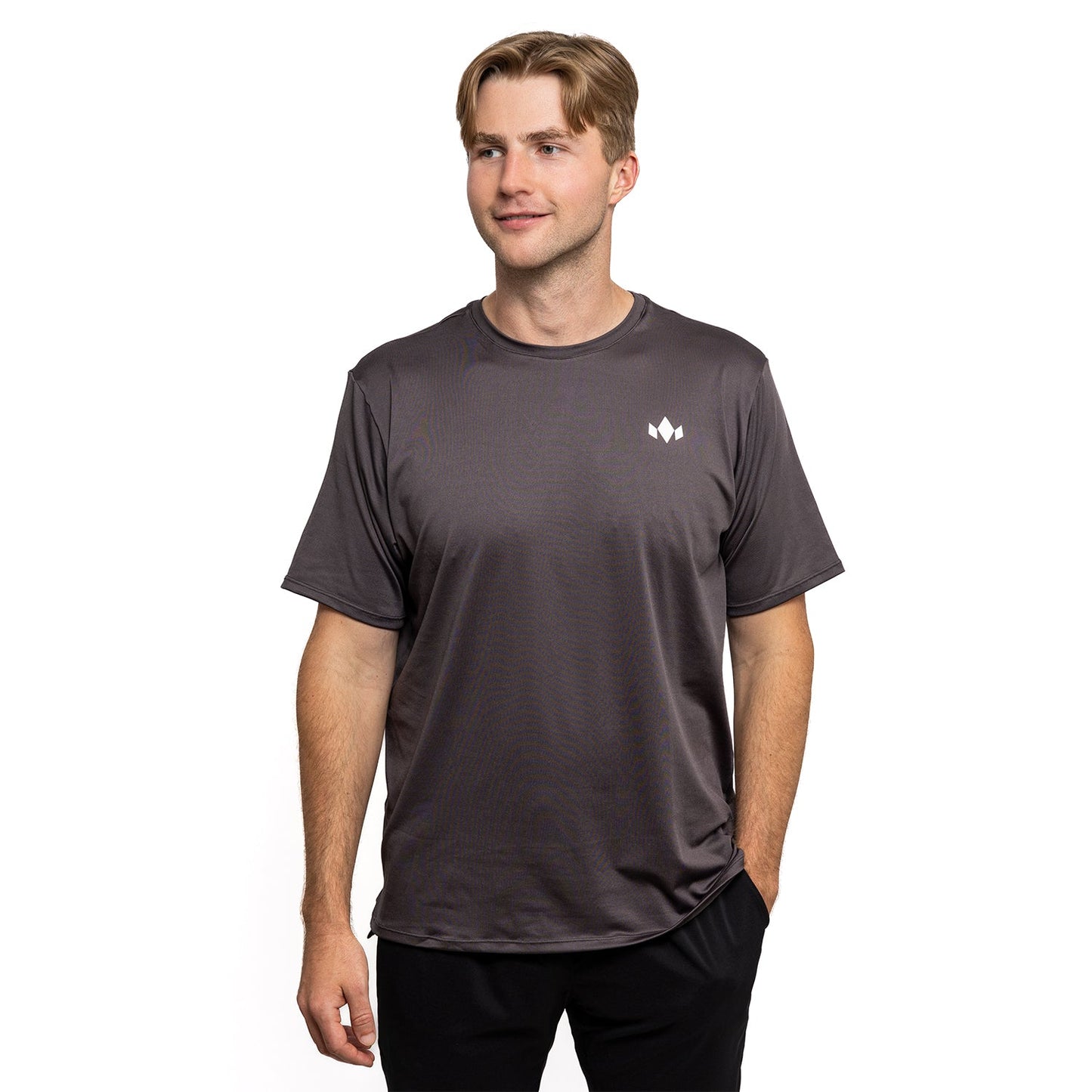 A young man with short light brown hair wears the Diadem Sports Men's Essential Crew Neck, a loose-fitting dark gray tee with a small white logo on the upper left chest. He stands facing forward, one hand in his pocket, smiling slightly against a white background.