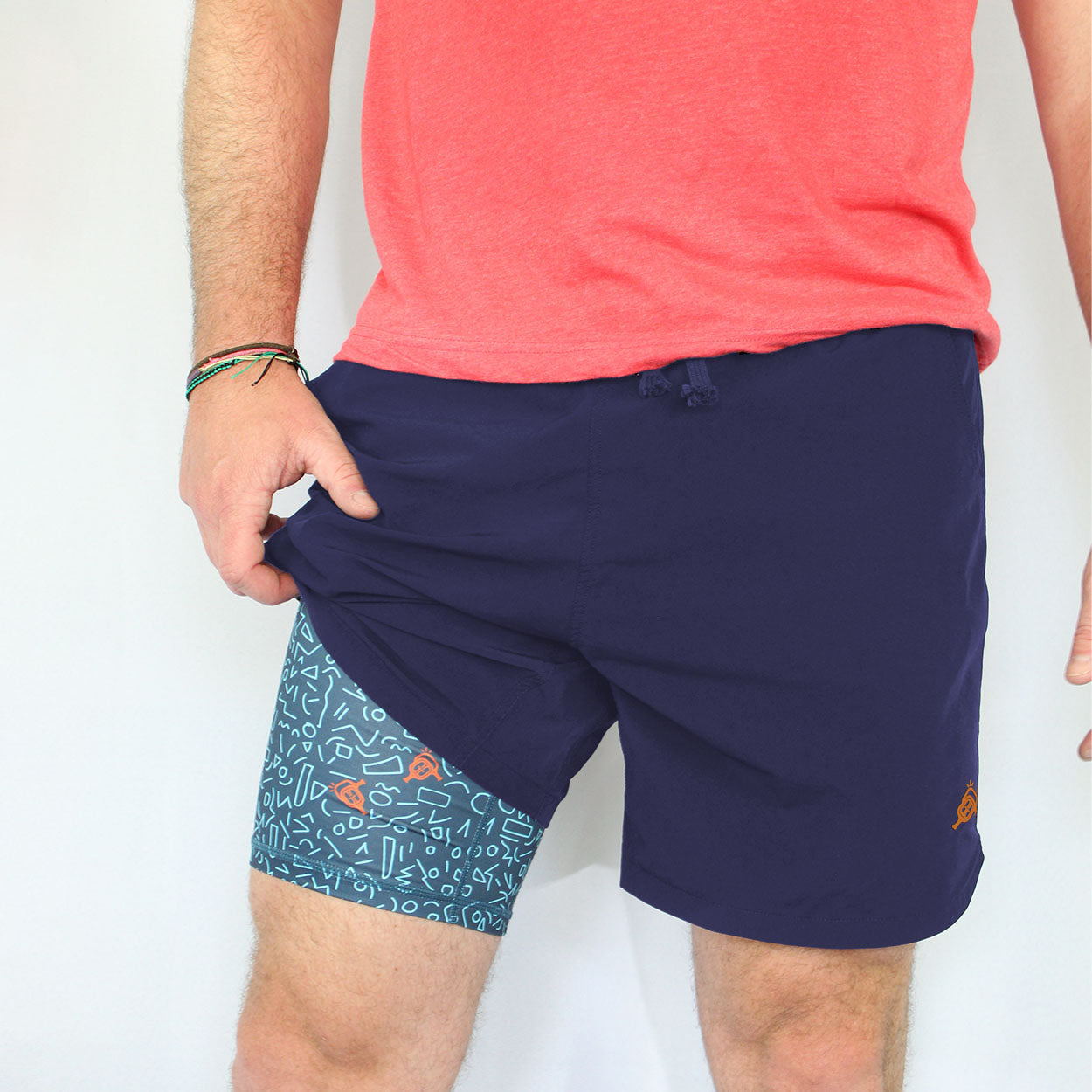 A person wearing a coral shirt and Good Get Pickleball Shorts, Court Clash Edition—navy blue with a blue panel featuring white doodle patterns—is shown pulling the shorts to reveal the unique design; only their torso and legs are visible.