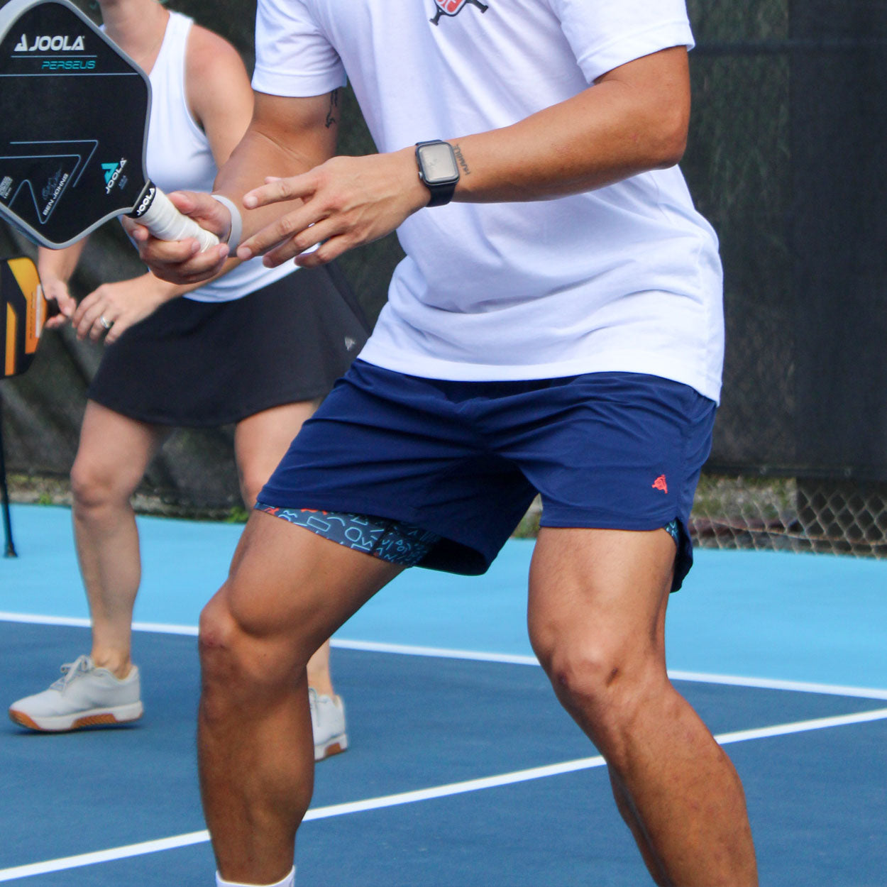 A man in Good Get Pickleball Shorts, Court Clash Edition, grips a paddle on the tennis court, ready for action.