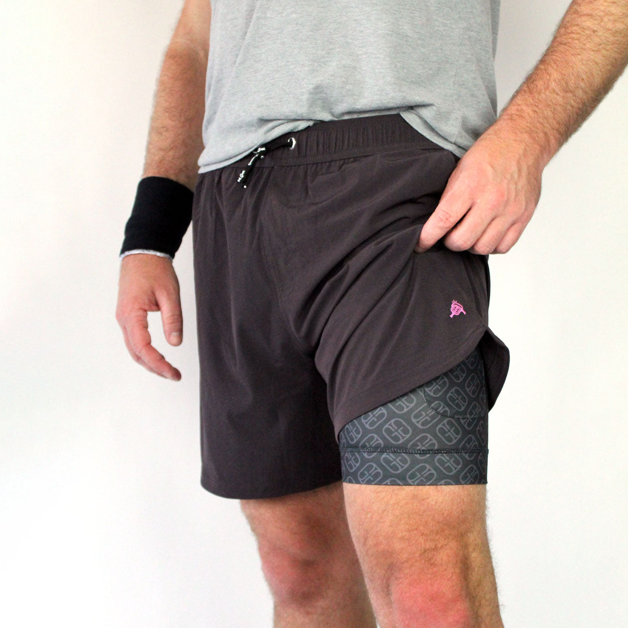 A close-up of someone in Good Get Pickleball Shorts, GG Edition—lifting the hem to reveal a compression liner beneath. A black wristband is on one arm; only their lower torso and upper legs are visible.