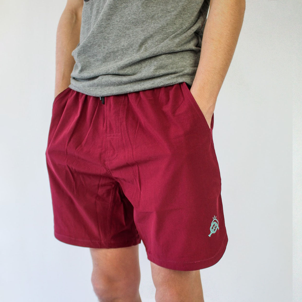 A person wears Good Get's Magenta Me Crazy Pickleball Shorts without liner in red and a grey t-shirt, standing with hands in pockets. The knee-length shorts have a small left hem logo, shown against a plain white background.
