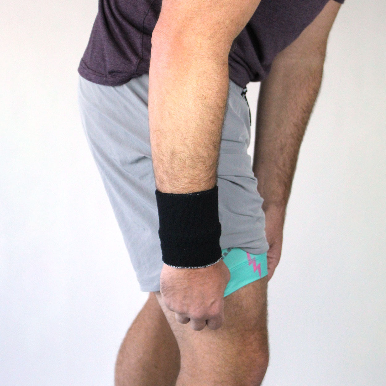 A person in a purple shirt and light gray Good Get Pickleball Shorts, PickleDome Edition, bends forward wearing a black wristband. They lift the shorts' hem to reveal light blue underwear with a pink zigzag pattern against a plain white background.