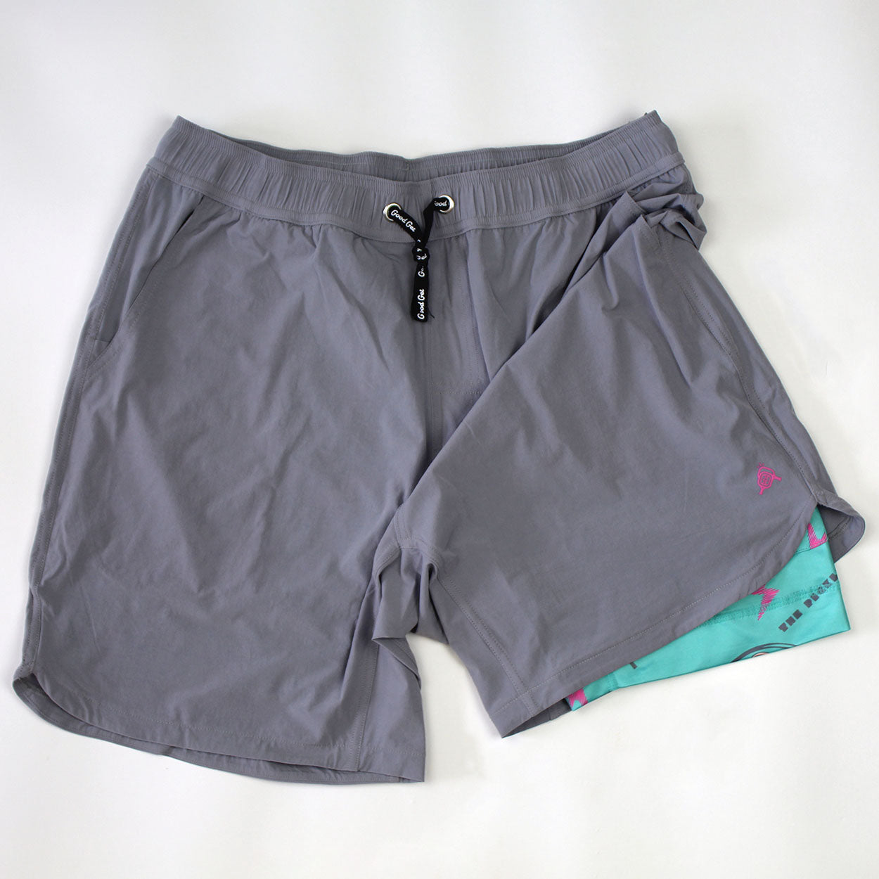 A pair of light gray Good Get Pickleball Shorts, PickleDome Edition, with elastic waistband, black drawstring, side pockets, turquoise PickleDome compression lining under the right leg, and a pink logo on the outer left hem.