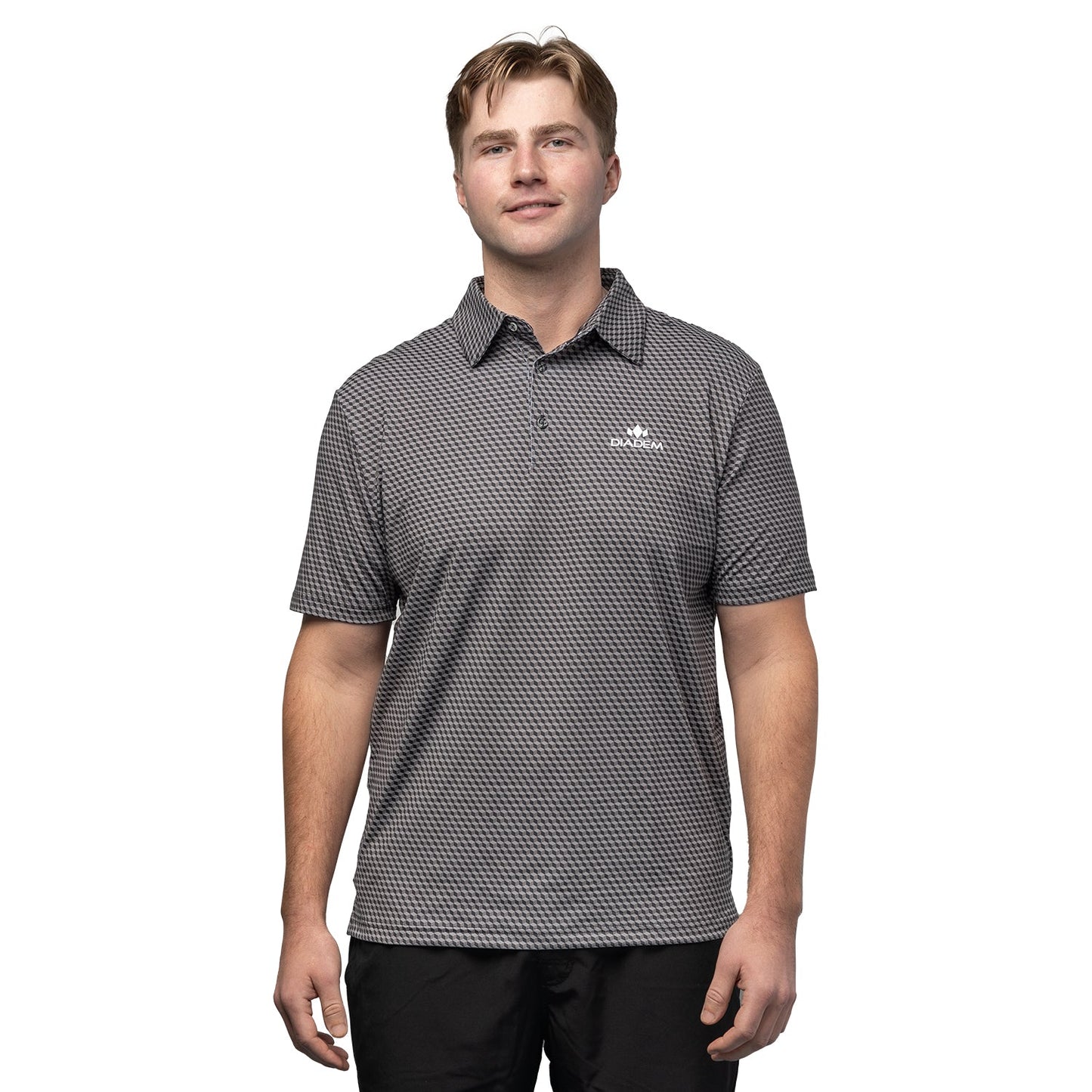 A young man with short, light brown hair and black pants faces forward, smiling slightly. He wears a Diadem Sports Men's Scales Competition 18K Pattern Polo from the Offshore Clubhouse Collection, featuring a small white logo on the chest.