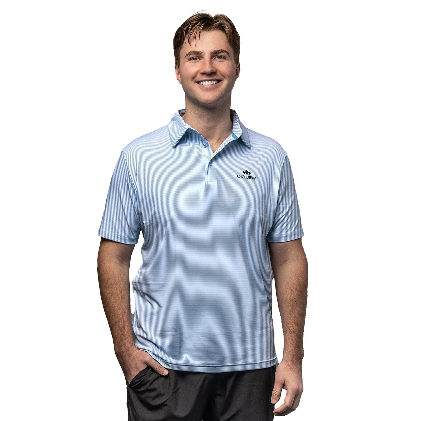 A smiling man with short brown hair wears the Men's Scales Weekender Striped Polo by Diadem Sports, featuring a small black "DAKOTA" logo and crown graphic on the chest. He stands against a plain white background with one hand in his pocket.