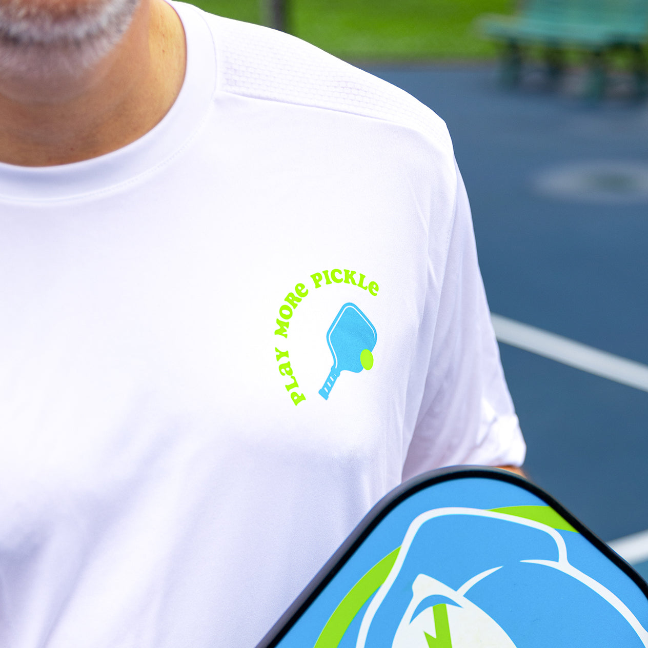 A man wearing the Swinton Pickleball Men's Pickled 'Em Performance Shirt in white, with a blue paddle graphic and "PLAY MORE PICKLE" text, stands on an outdoor court holding a colorful paddle; blurred benches and green fence are in the background.