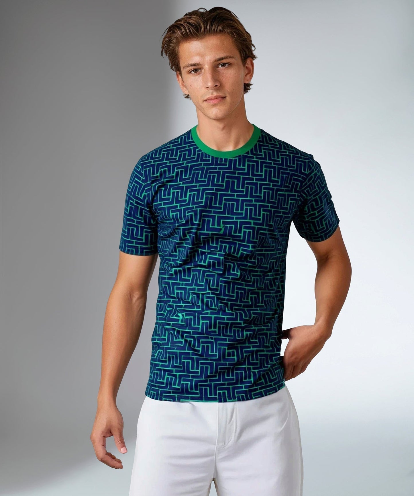 A young man with light brown hair and a neutral expression stands against a plain background wearing inPhorm NYC's Men's Short-Sleeve Crew Neck—UV Protection, Antimicrobial—in blue with a green geometric maze pattern, paired with white pants.