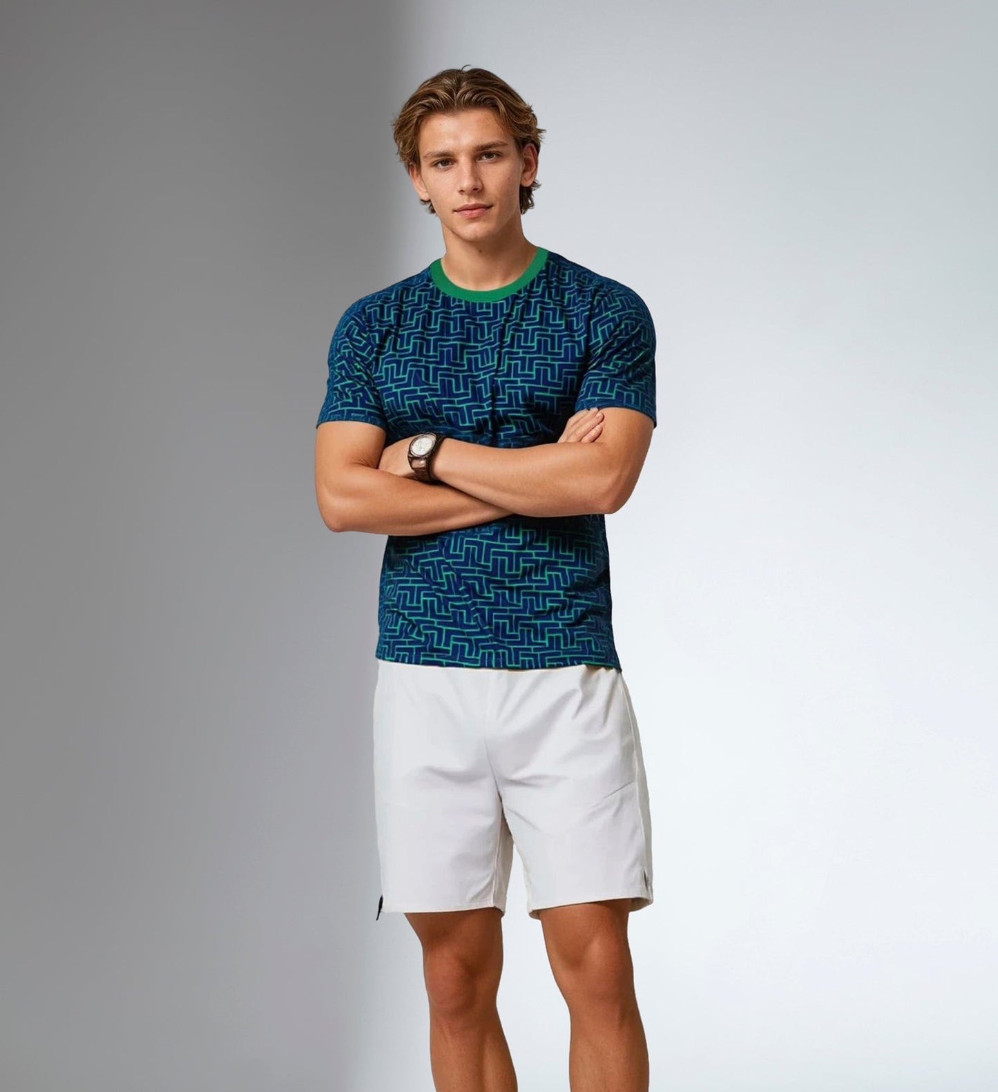 A young man with short light brown hair stands with arms crossed, wearing an inPhorm NYC Men's Short-Sleeve Crew Neck—UV Protection, Antimicrobial—and white shorts. He sports a watch on his left wrist against a softly lit light gray background.