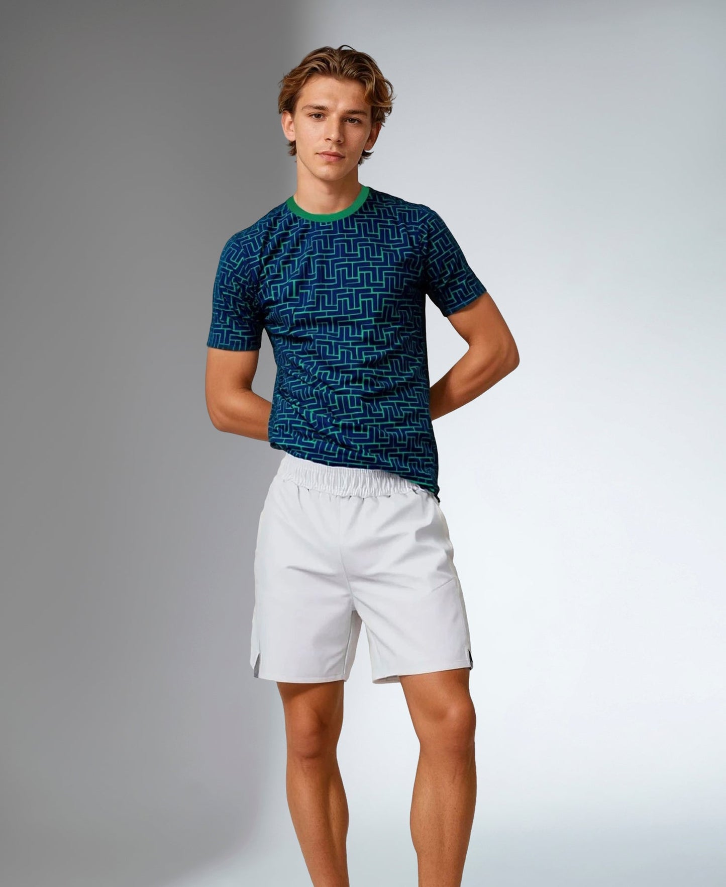 A young man with light brown hair stands against a plain gray background, wearing inPhorm NYC's Men's Short-Sleeve Crew Neck with UV protection and antimicrobial features, paired with white athletic shorts. His hands rest behind his back as he gazes calmly at the camera.