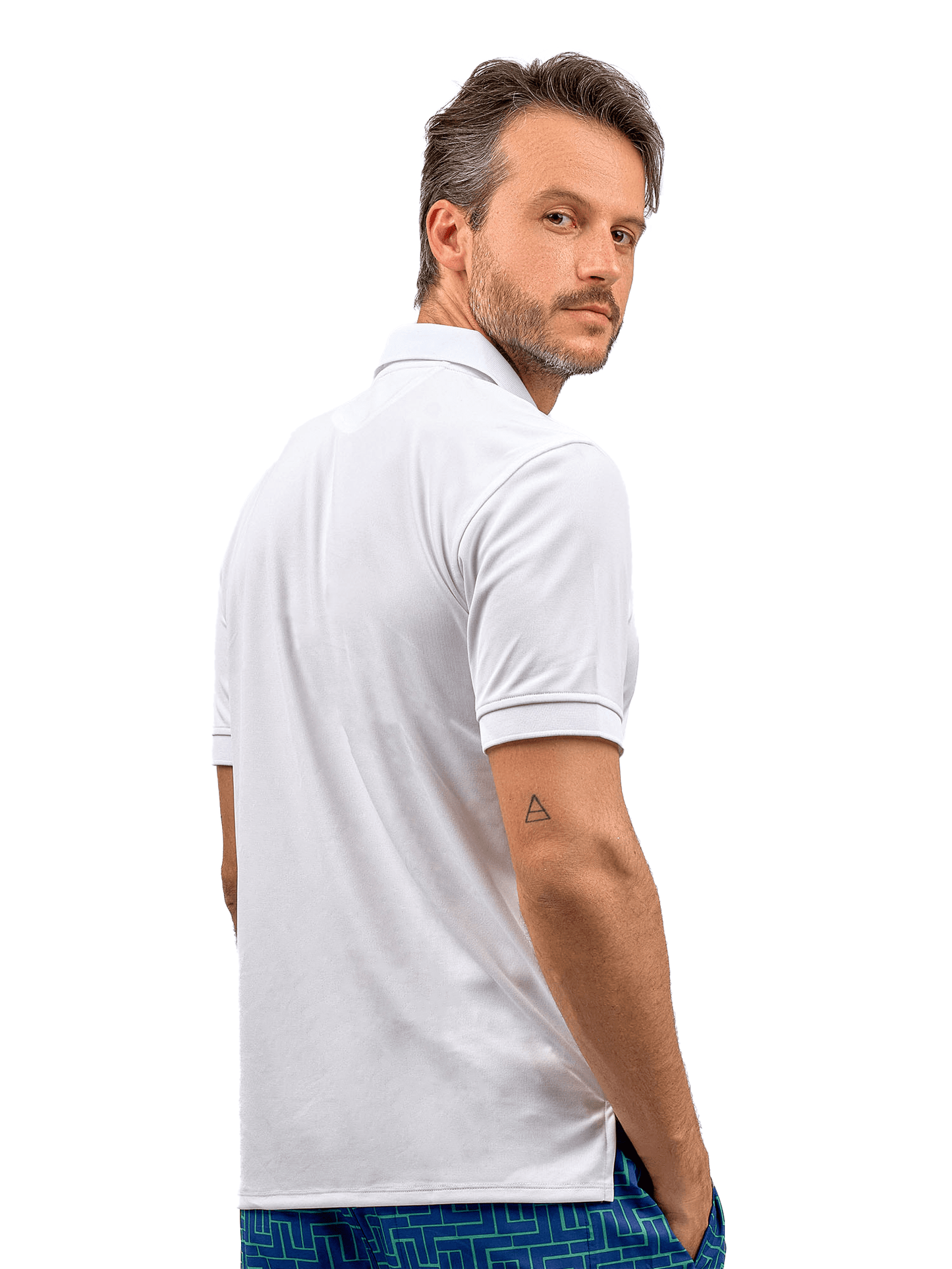 A man with short brown hair and light stubble stands in a three-quarter pose wearing the inPhorm NYC Men's Spin Polo shirt, a performance shirt for tennis & golf, paired with blue patterned shorts. His right hand is in his pocket, showing a triangle tattoo.