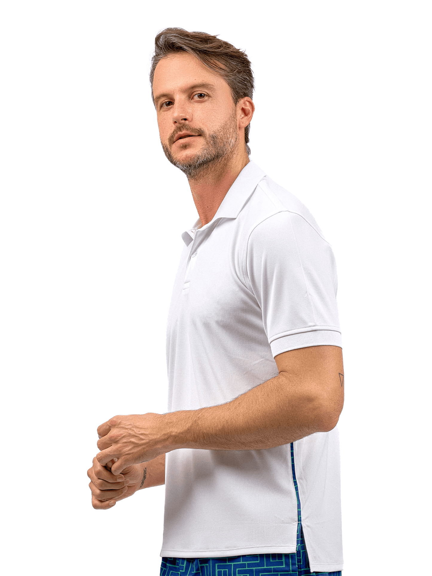 A man with short brown hair and a trimmed beard wears the inPhorm NYC Men's Spin Polo shirt—Performance Shirt for Tennis & Golf—in white, paired with blue patterned shorts. He stands against a white background, facing slightly to the side, hands loosely clasped.
