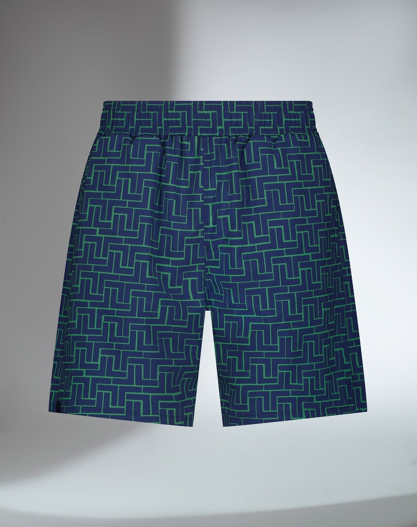 The inPhorm NYC Men's Spin Tennis Shorts - Tessel, in dark blue with a green geometric pattern, feature an elastic waistband, pockets, and UV protection. Displayed on a softly lit neutral background with a subtle shadow on the left.