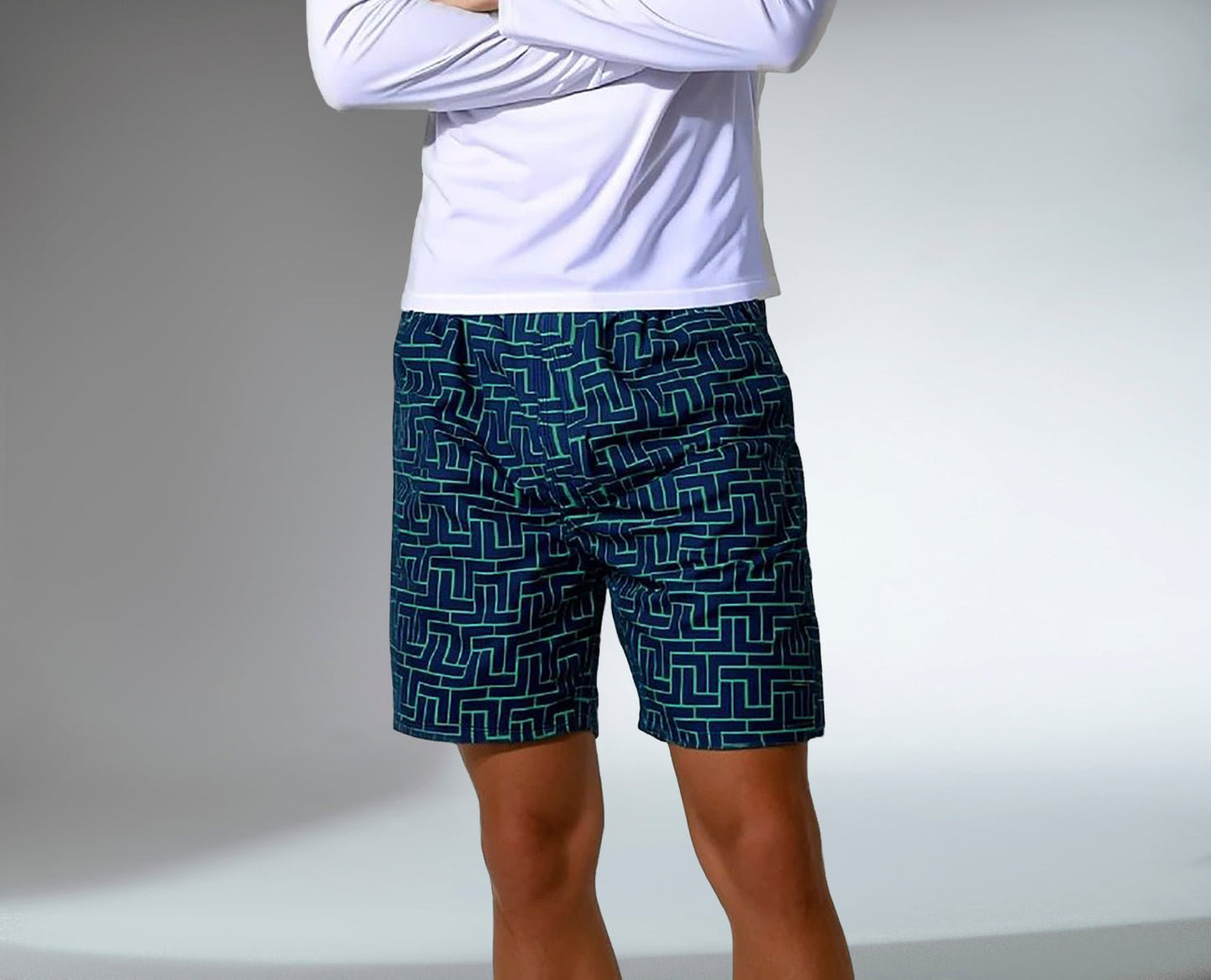 A person stands with arms crossed, wearing a long-sleeve white shirt and inPhorm NYC Men's Tessel Spin Tennis Shorts—dark blue athletic shorts with UV protection, pockets, and a light blue and green geometric maze pattern. The softly lit background is neutral.