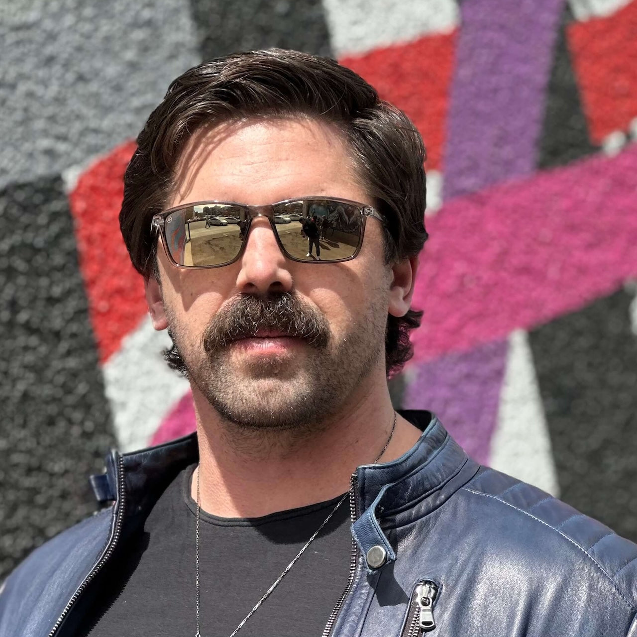A man with brown hair and a mustache wears Epoch Eyewear’s Murphy polarized sunglasses, a blue leather jacket, and a black shirt as he stands in bright sunlight before a mural of bold geometric shapes in black, white, red, and purple.