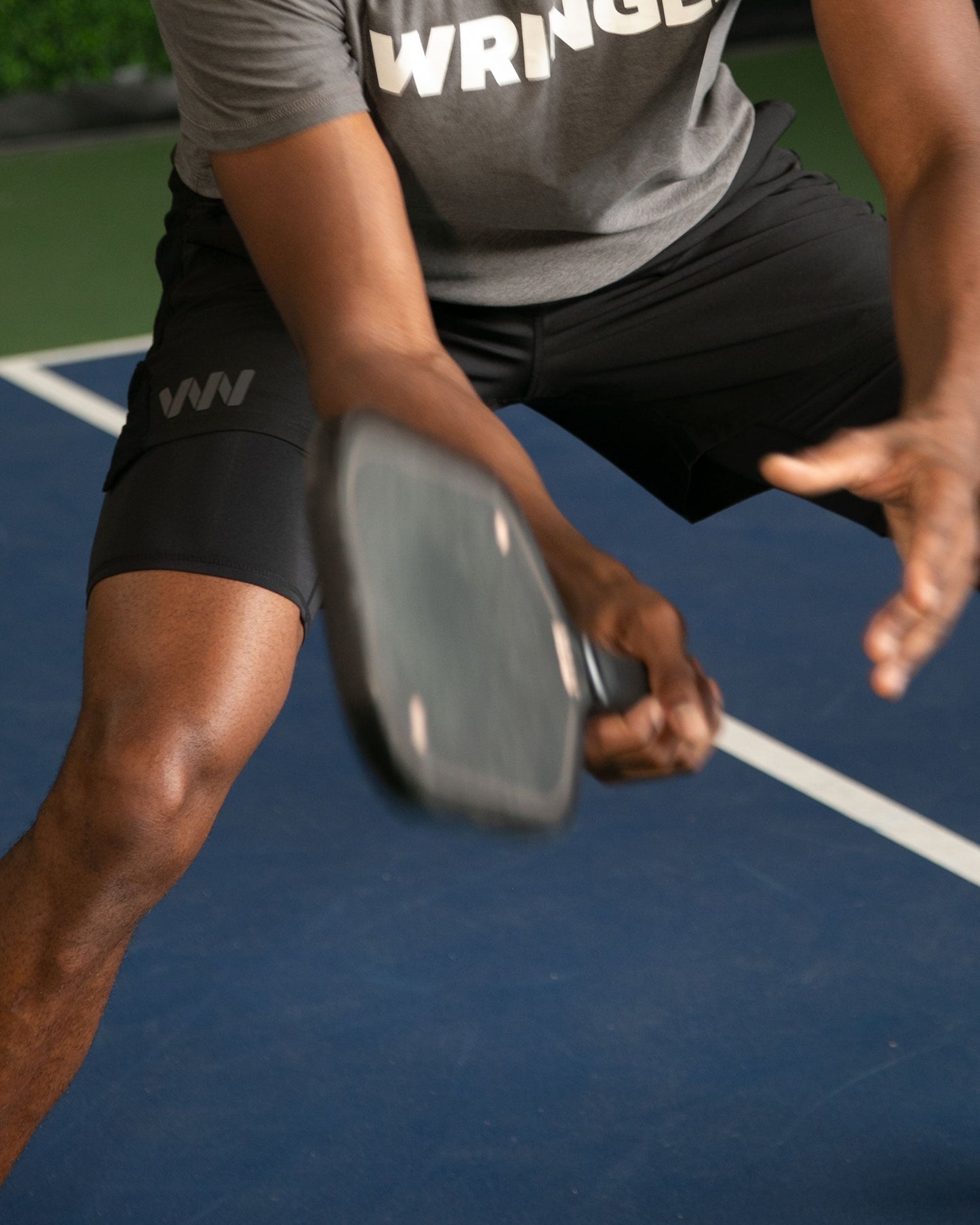 Wearing Wringer Wear's Mojave Performance Shorts 6" Lined and a gray moisture-wicking shirt with partial white text, a person bends their knees on a blue pickleball court, focused on hitting the ball with a black paddle. Only torso and limbs are visible.