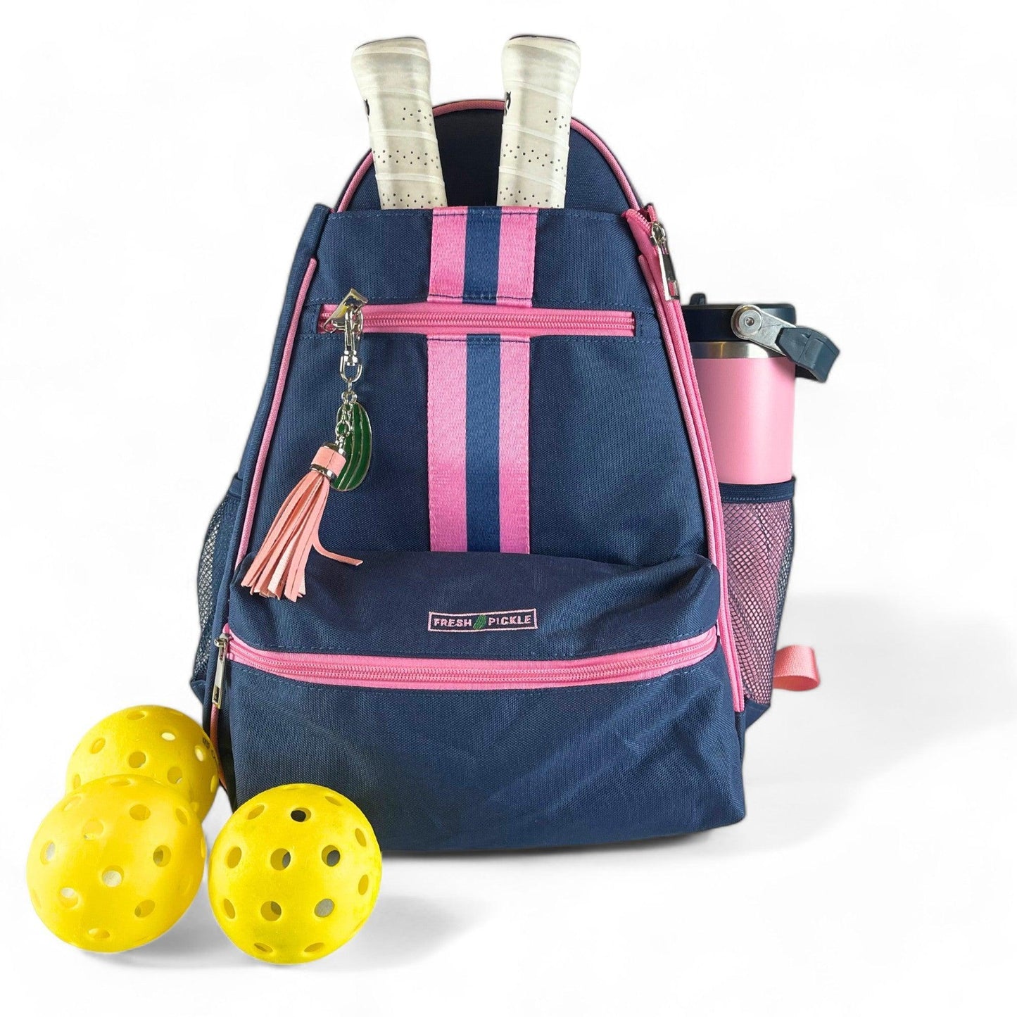 The Fresh Pickle Teddi Pickleball Backpack in navy with pink zippers and stripes holds two white-handled paddles, a pink water bottle, and a keychain with tassel and charm. Three yellow pickleballs sit in front of this hands-free bag.