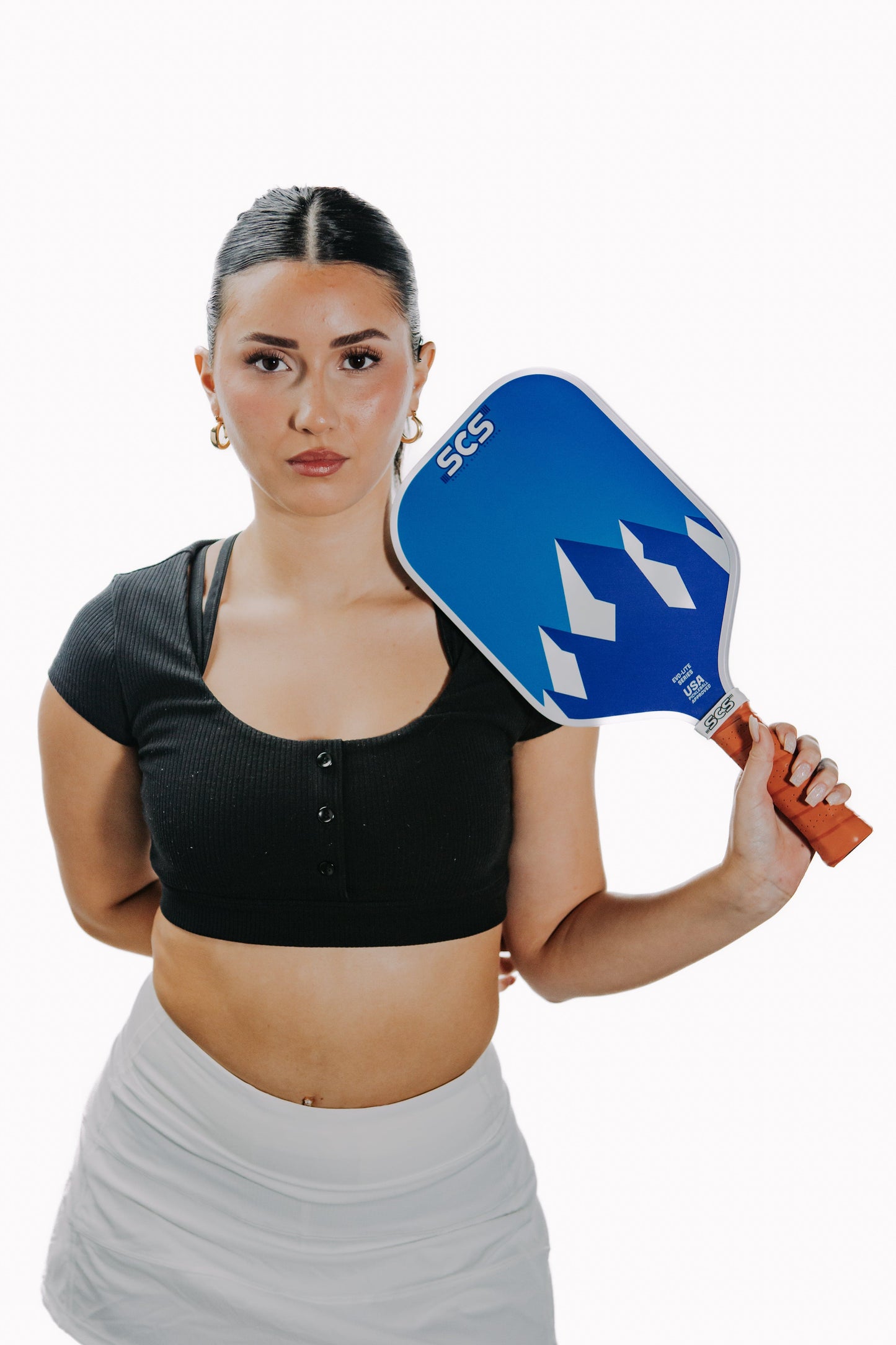 A young woman with dark hair pulled back, wearing a black crop top and white skirt, stands against a white background holding the Salted City Sports New Skool Evo-Lite Series pickleball paddle with a geometric design on her shoulder.