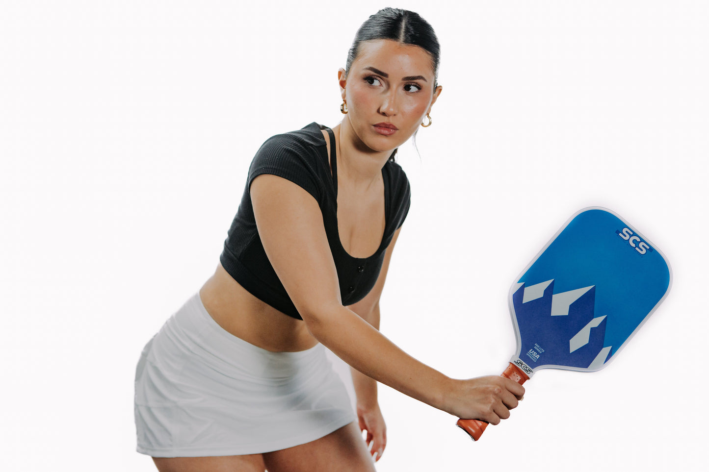 A woman with dark hair in a ponytail, wearing a black crop top and white skirt, holds the Salted City Sports New Skool Evo-Lite Series pickleball paddle in an athletic stance. USA Pickleball approved. Plain white background.