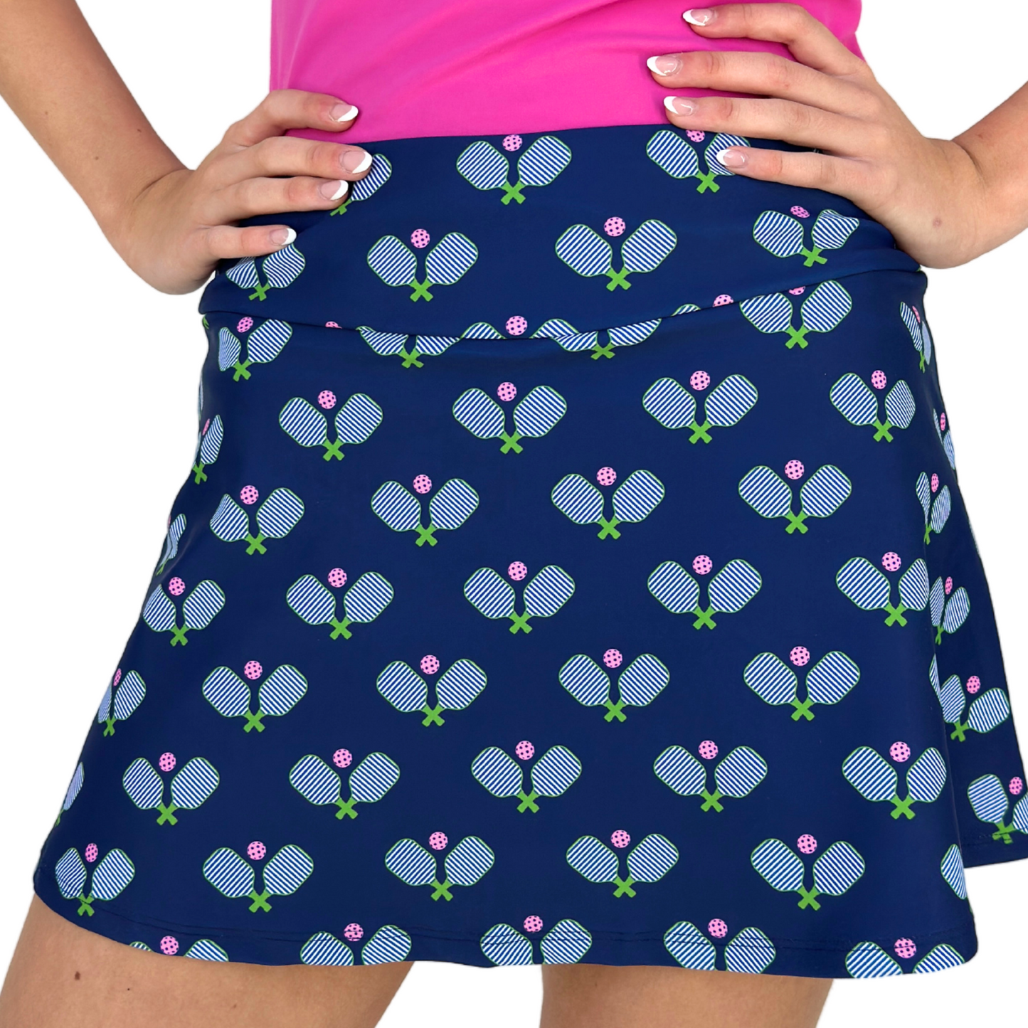 A person wears the Queen of the Court Hit It! Pickleball Skirt—navy blue, USA-made, with crossed paddles and pink pickleballs—paired with a bright pink top. Pink nails complete the look, matching both the activewear and playful skirt pattern.