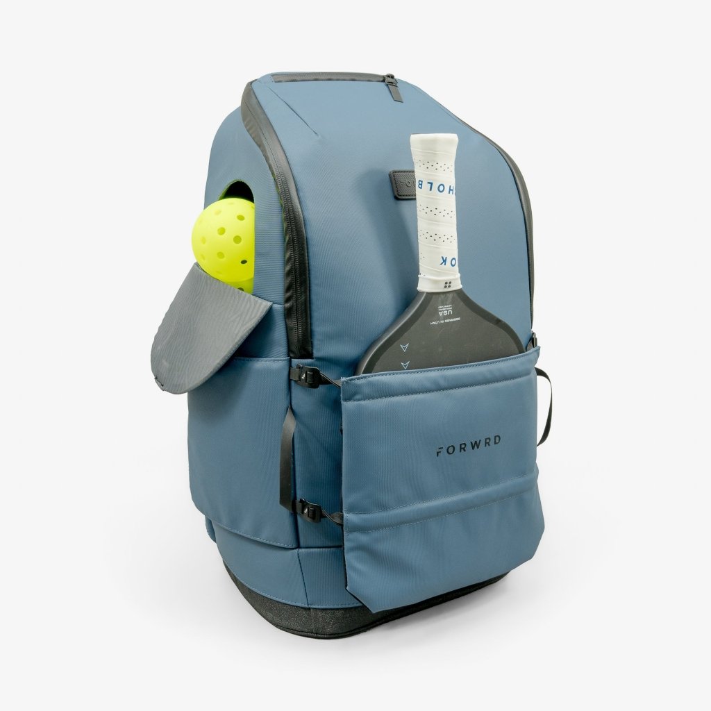 The FORWRD Court Caddy Pickleball Bag, in blue with black accents, stands upright on a white background. It features a front pocket holding a paddle, a tennis ball in the side pocket, and displays the brand’s name on the front.