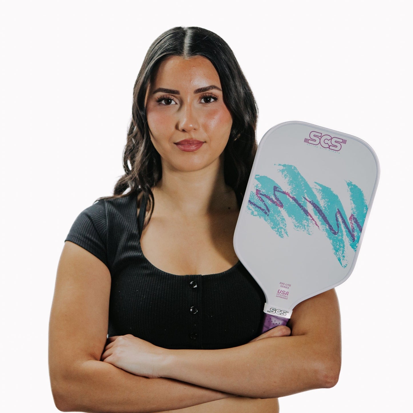 A confident woman with long dark hair in a black top stands arms crossed, holding the Salted City Sports Nostalgia | Evo-Lite Series pickleball paddle, which features teal and purple designs and "SCS" lettering, against a white background.