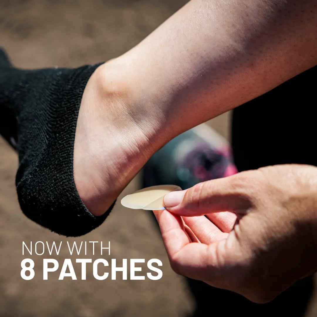A person peels a KT Health Blister Treatment Patch by KT Tape to apply on their heel, sock partially down. The background is blurred. White text at bottom left reads, “NOW WITH 8 PATCHES.”.