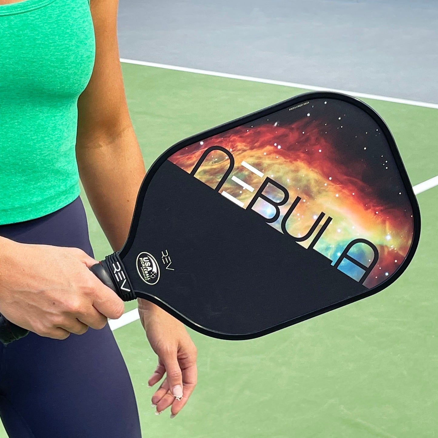 A person in a green tank top and dark pants holds the REV Pickleball NEBULA Pickleball Paddle, featuring a cosmic design, honeycomb core, black handle, and "NEBULA" branding, on a green outdoor court.